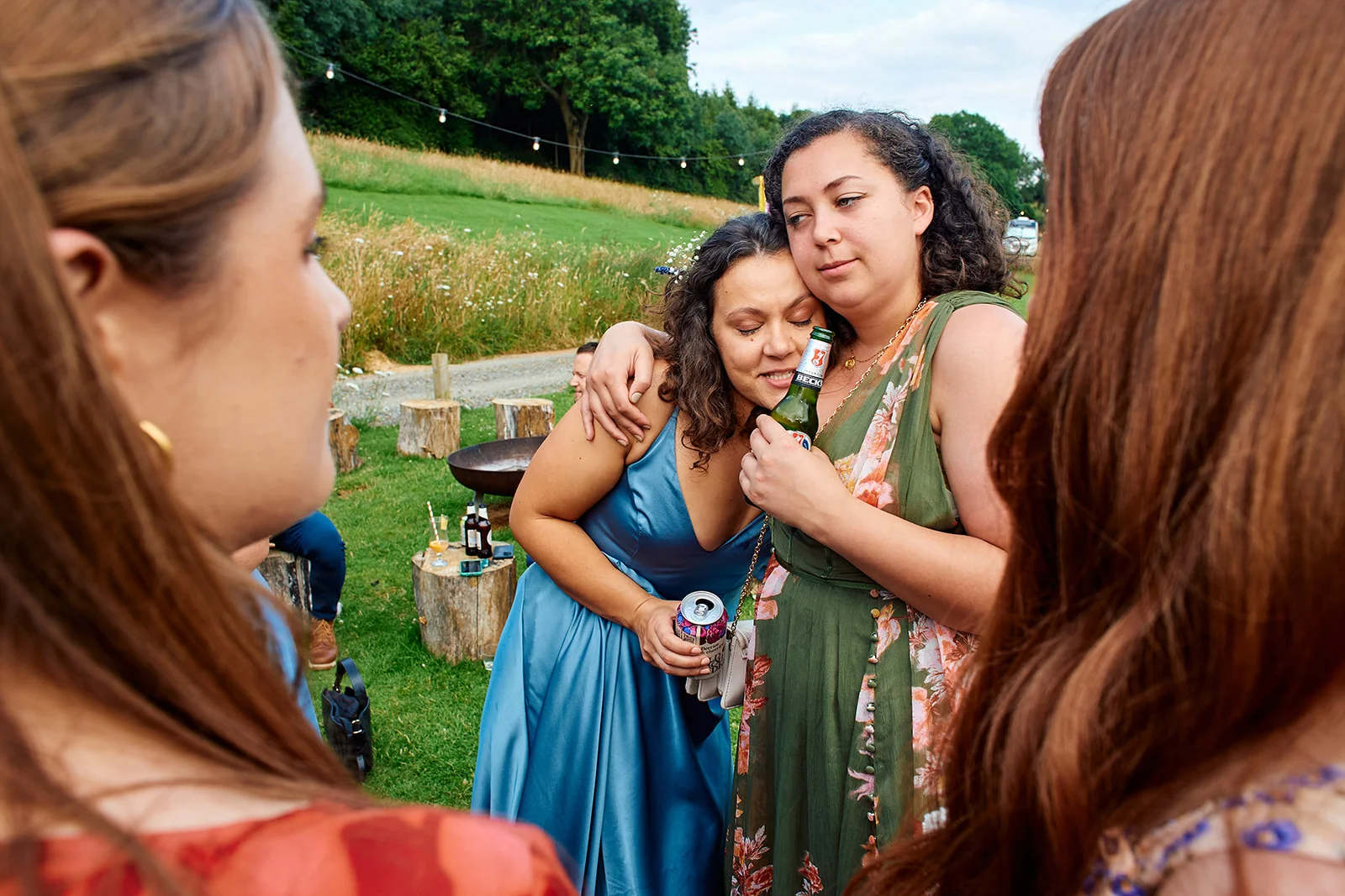 Two wedding guests share a warm embrace outdoors at a festival-style reception while holding drinks, framed by other guests in the foreground.