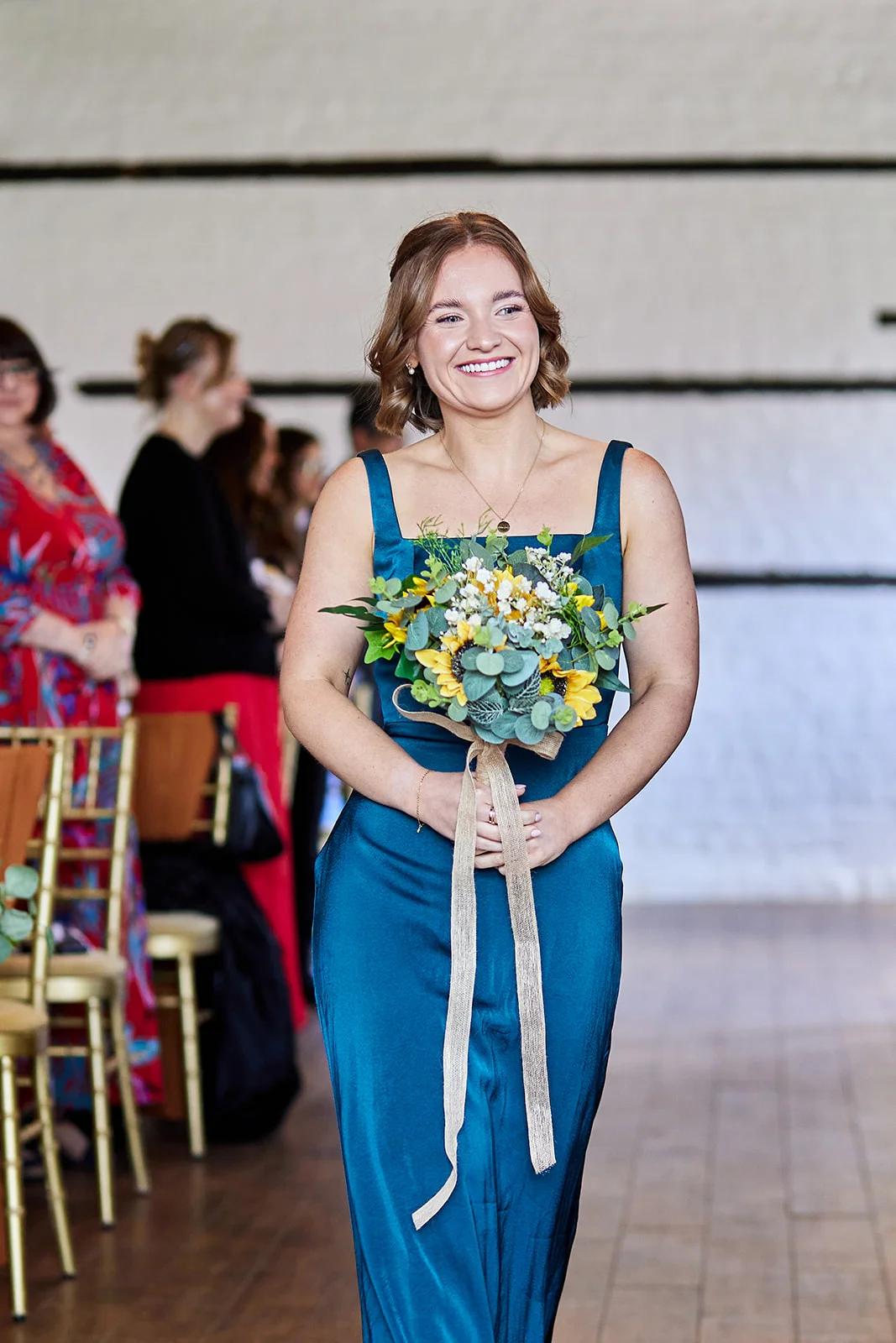 A smiling bridesmaid in a teal satin gown walks down the aisle holding a bouquet of sunflowers and greenery inside a venue with exposed brick walls.