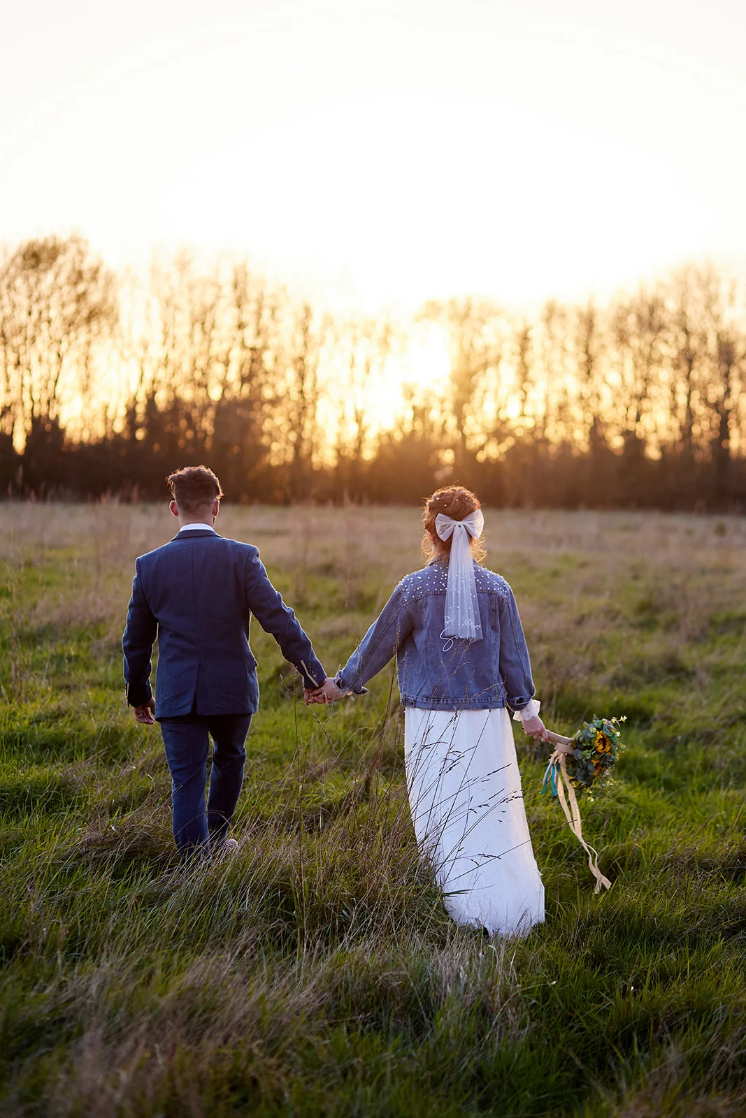 Newlyweds walk hand in hand through a grassy field during golden hour, with the bride wearing a denim jacket and holding a wildflower bouquet.