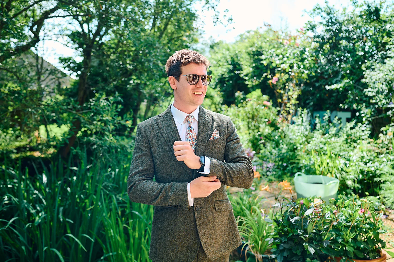 A smiling groom wearing a tweed suit, patterned tie, and sunglasses stands in a lush garden setting while adjusting his cuff.