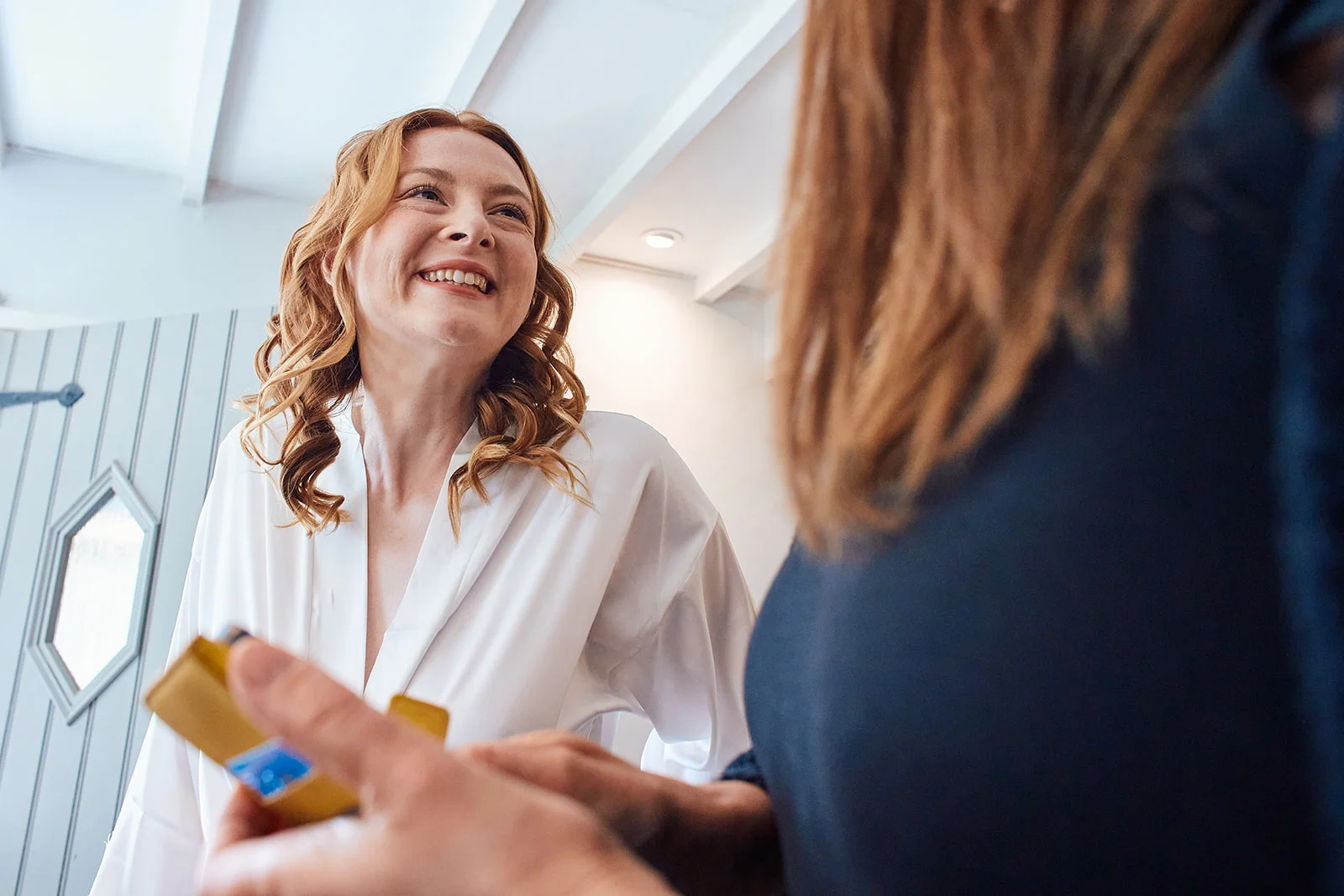 A smiling bride with red curly hair wearing a white getting ready robe looks happily towards someone holding a yellow item in the foreground.