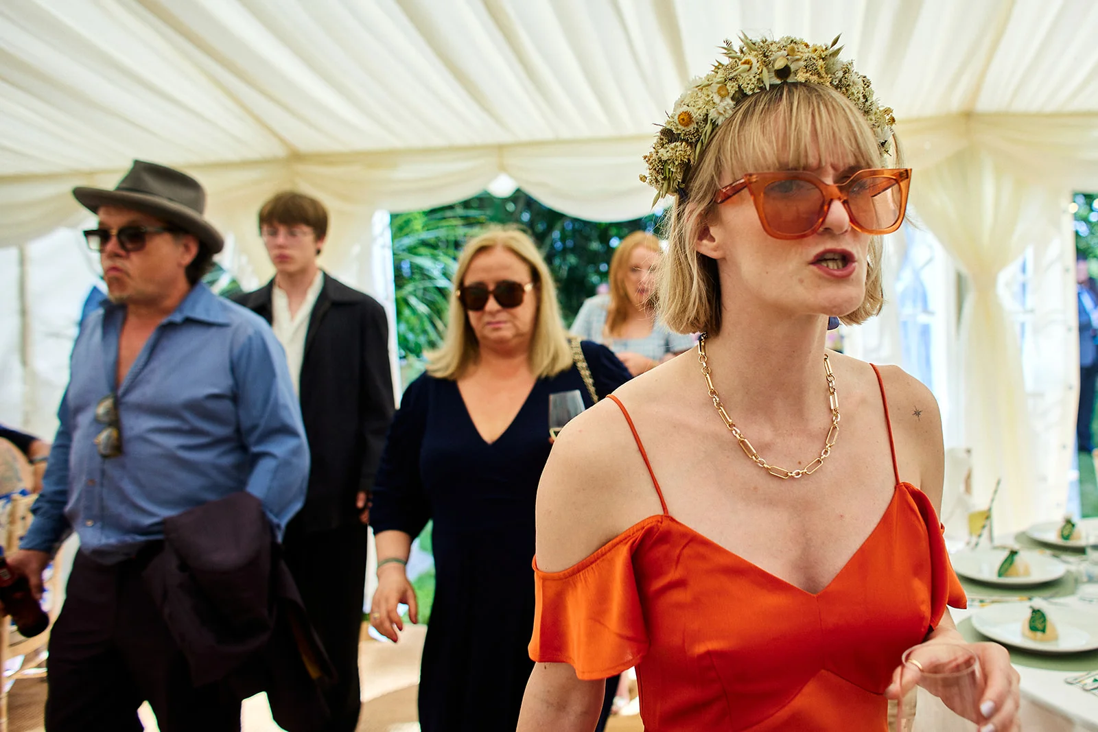 A candid shot of wedding guests mingling inside a marquee, featuring a woman in the foreground wearing an orange dress and floral crown.