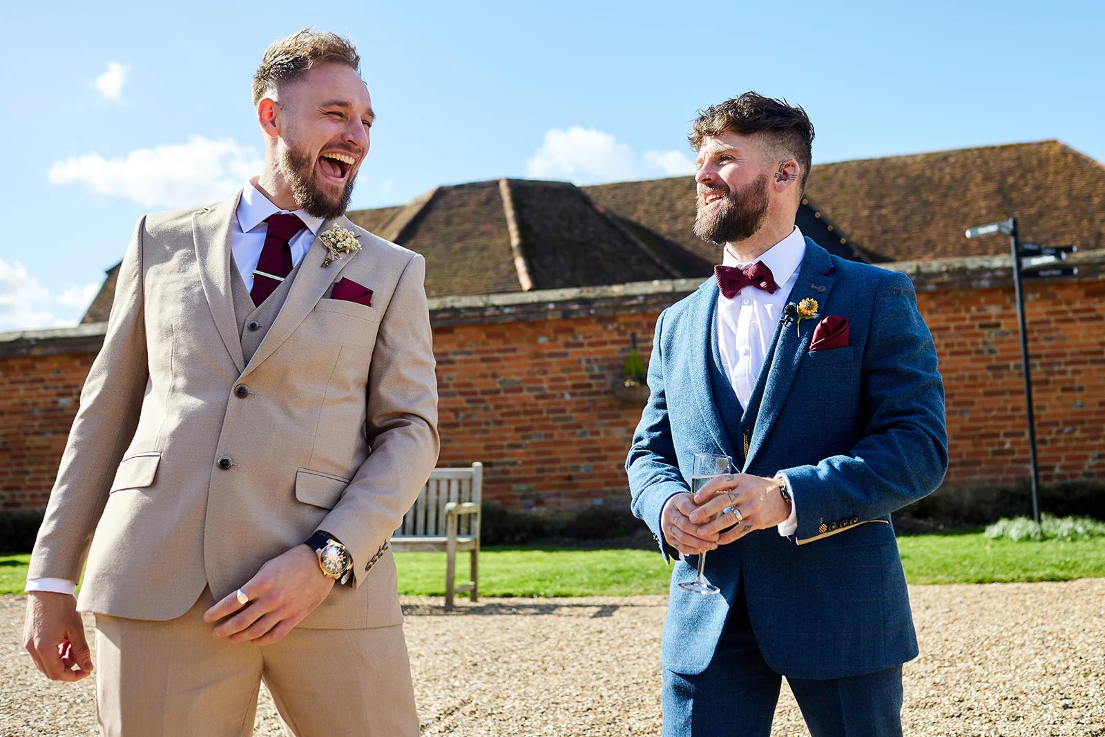 Two grooms in colourful suits laugh together outside a rustic stone and brick barn on a sunny wedding day.
