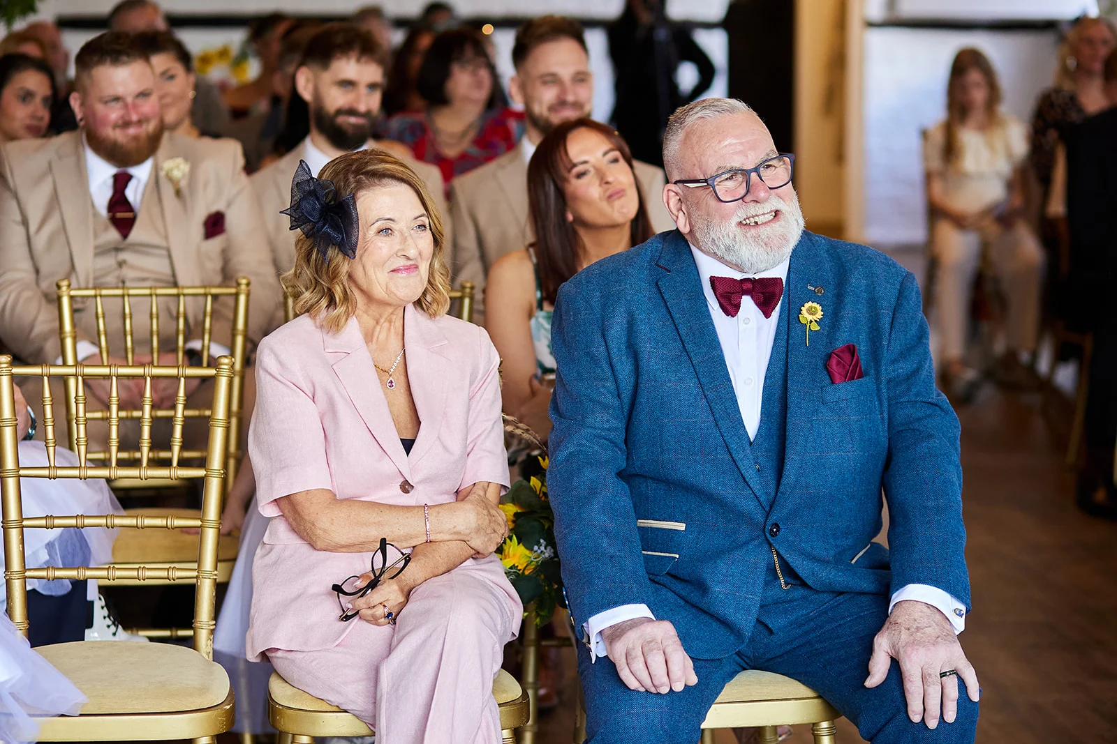 An older couple seated proudly in the front row of a wedding ceremony, with the smiling father wearing a blue suit and the mother in a pink trouser suit.
