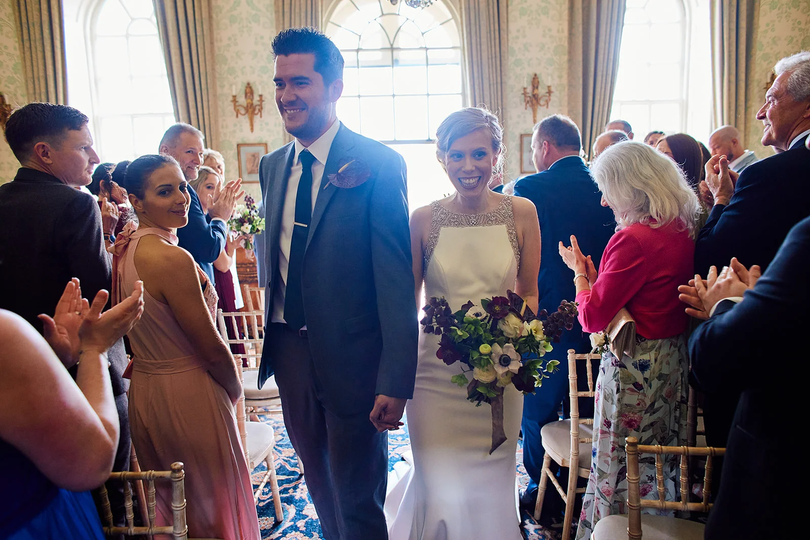 Newlyweds walk back down the aisle hand in hand, smiling joyfully as guests applaud them in a brightly lit, elegant venue.