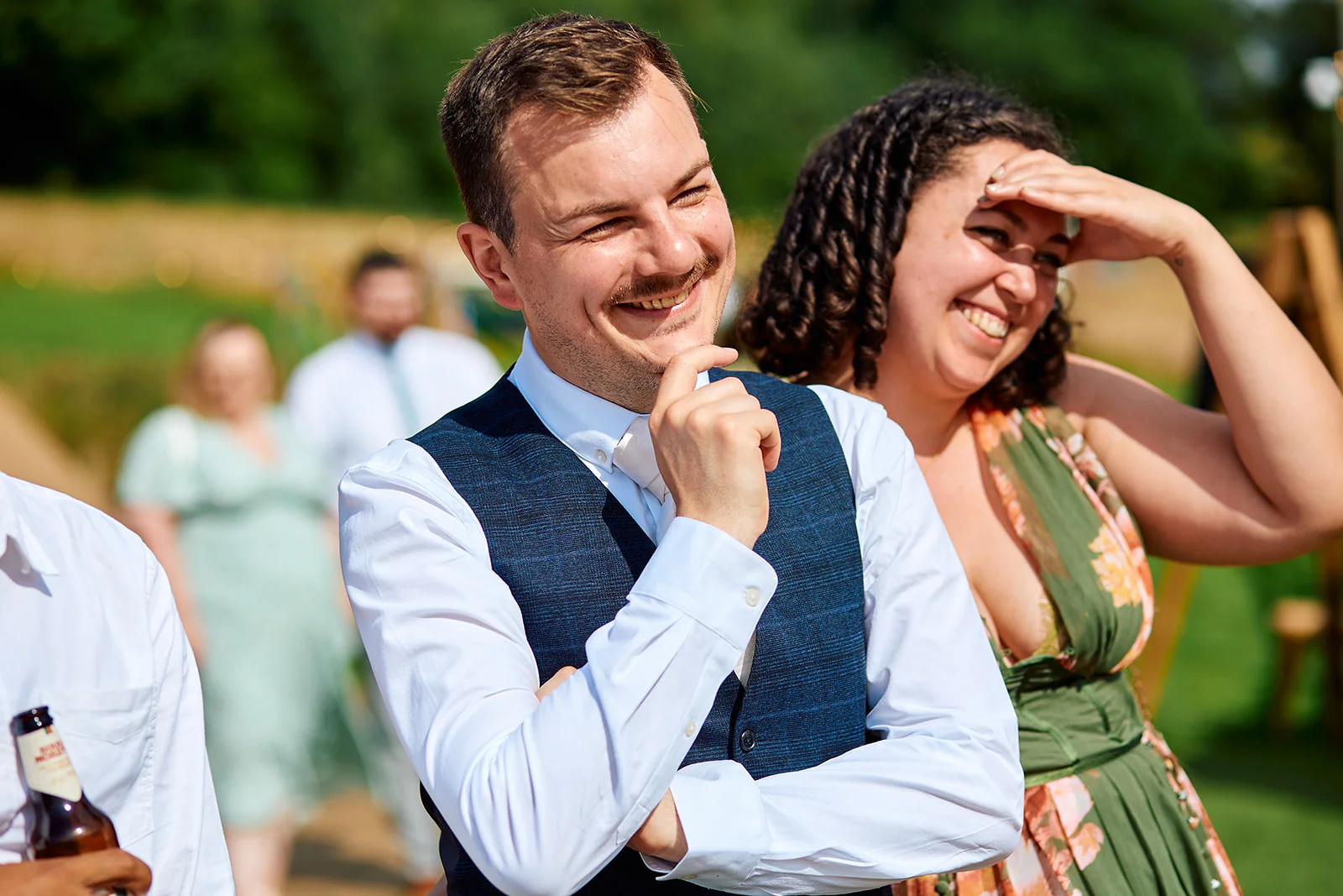 A smiling groom wearing a blue tweed waistcoat and white shirt laughs outdoors on a sunny wedding day, accompanied by guests in the background.