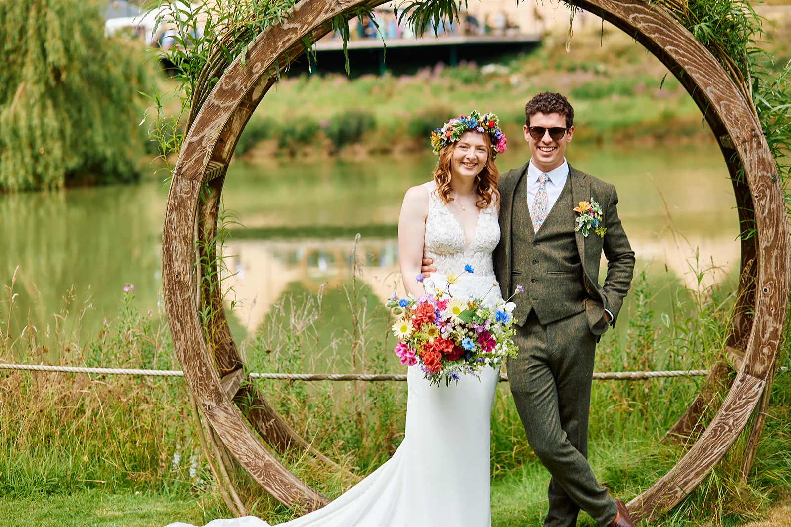 A smiling bride and groom pose together outdoors next to a lake, framed by a large circular wooden arch adorned with foliage.