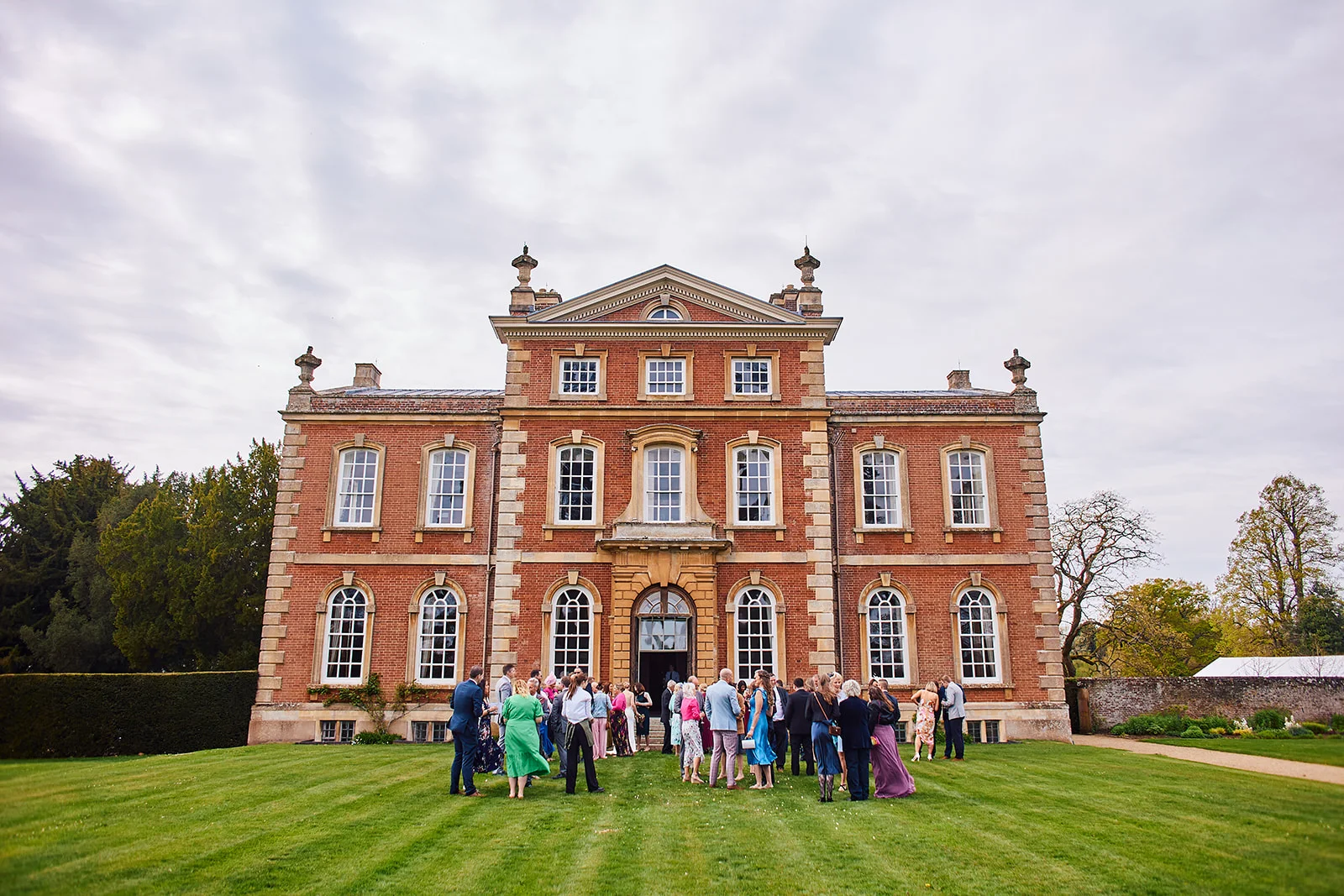 Wedding guests mingle on the front lawn of a grand red brick country house under an overcast sky during the drinks reception.