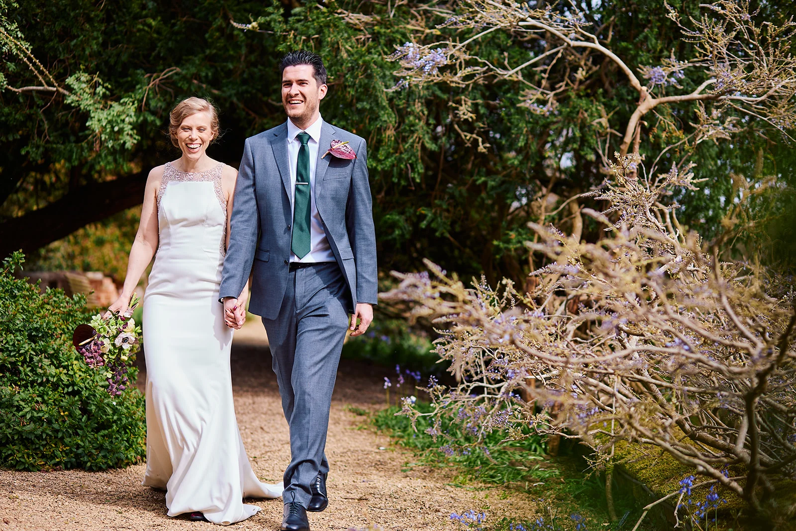 A joyful bride and groom walk hand in hand along a gravel path surrounded by lush greenery and flowering shrubs, both smiling broadly in the sunshine.