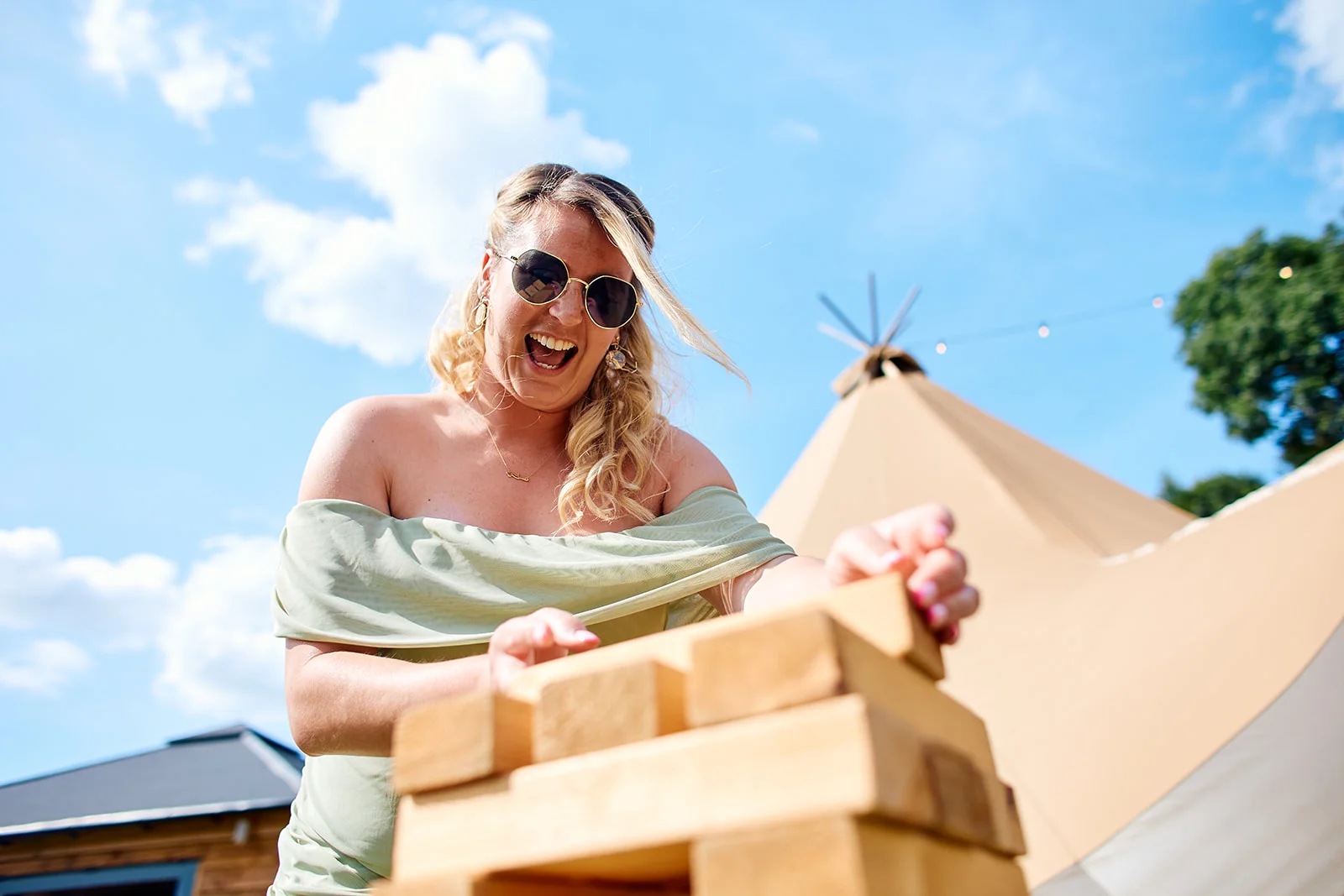 A smiling guest wearing sunglasses and an off-the-shoulder sage dress plays giant jenga outside during a festival-style tipi wedding on a sunny day.