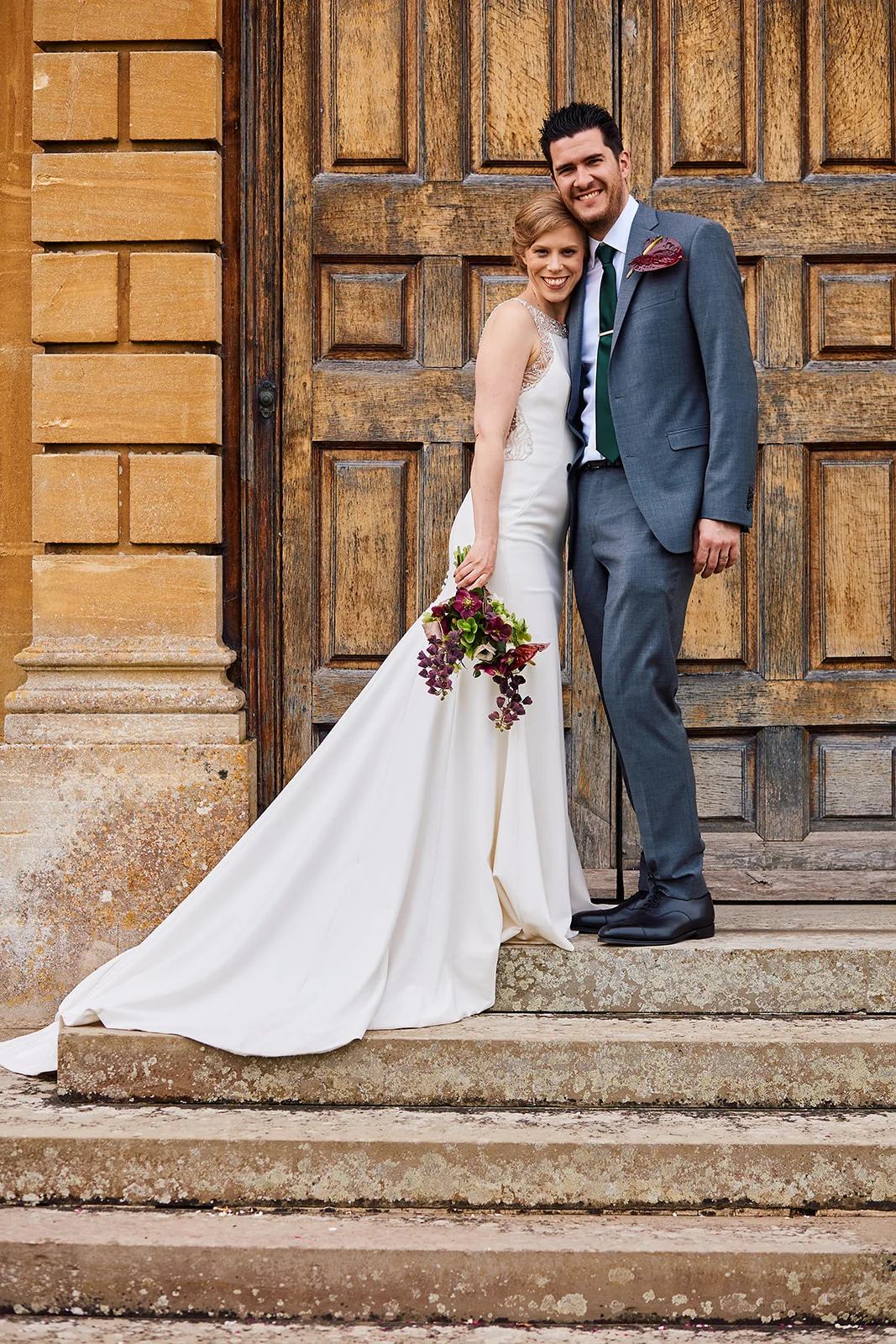 A smiling bride holding a cascading bouquet and groom in a grey suit stand embracing on stone steps in front of a large wooden door.