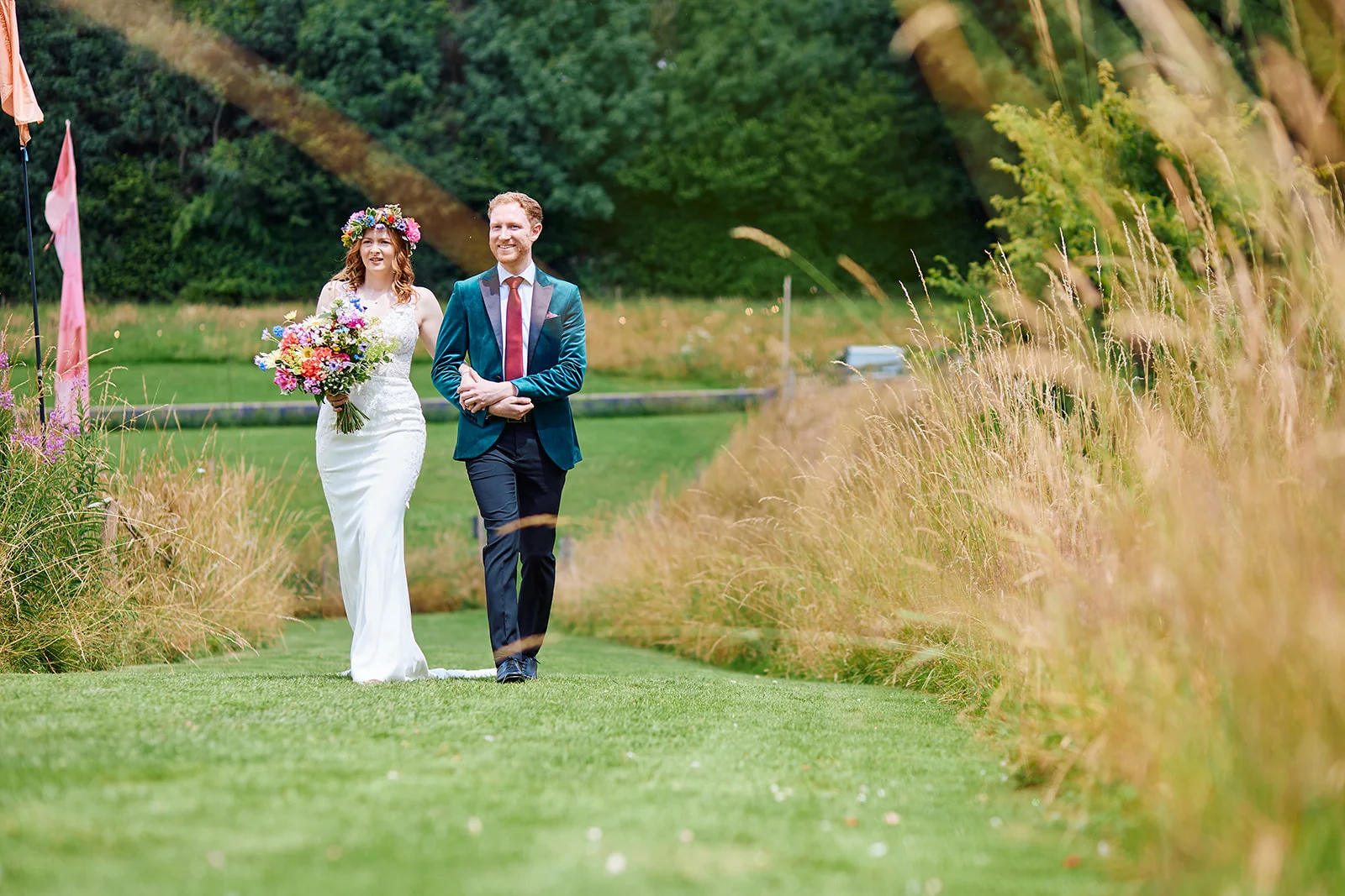 A newlywed couple walk hand-in-hand along a grassy path flanked by tall wildflowers, with the bride wearing a flower crown and carrying a colourful bouquet while the groom wears a velvet dinner jacket.