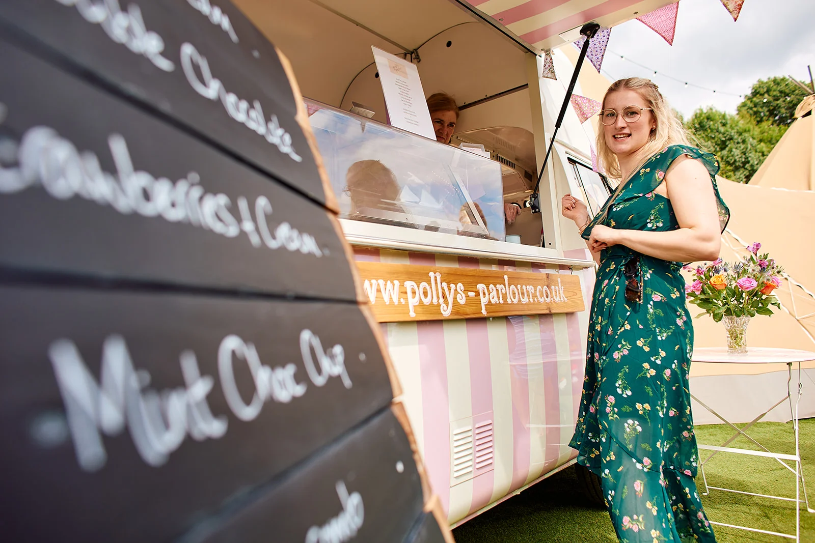 A wedding guest in a green floral dress poses next to a pink and white striped ice cream van during an outdoor festival-style reception.