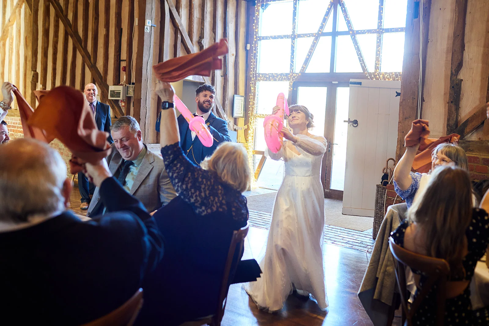 A joyful bride enters a rustic oak-framed barn wedding reception while cheering guests wave napkins and inflatable sticks in the air.