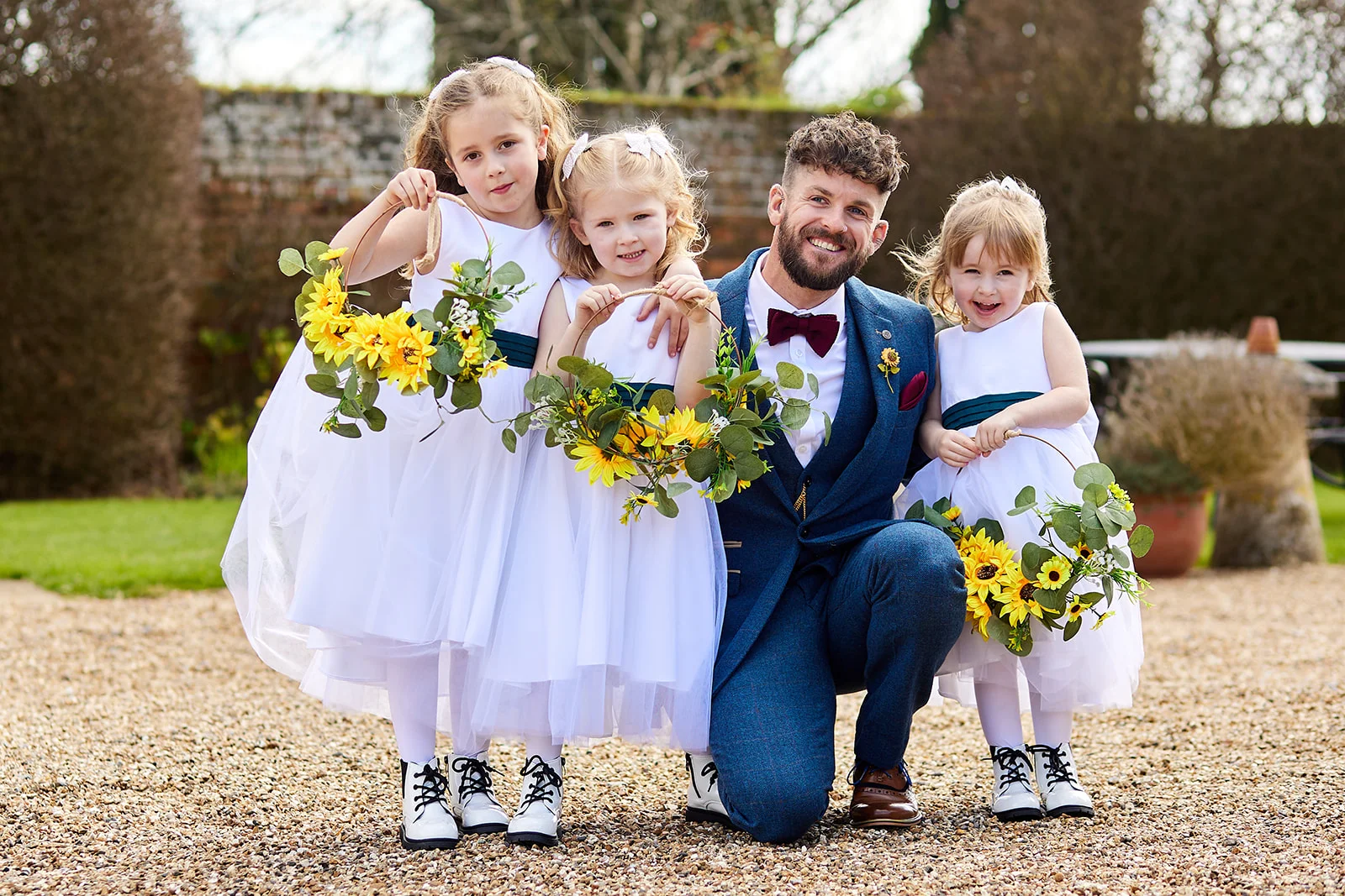 A smiling groom in a blue tweed suit poses outdoors with three young flower girls holding floral hoops adorned with yellow sunflowers.