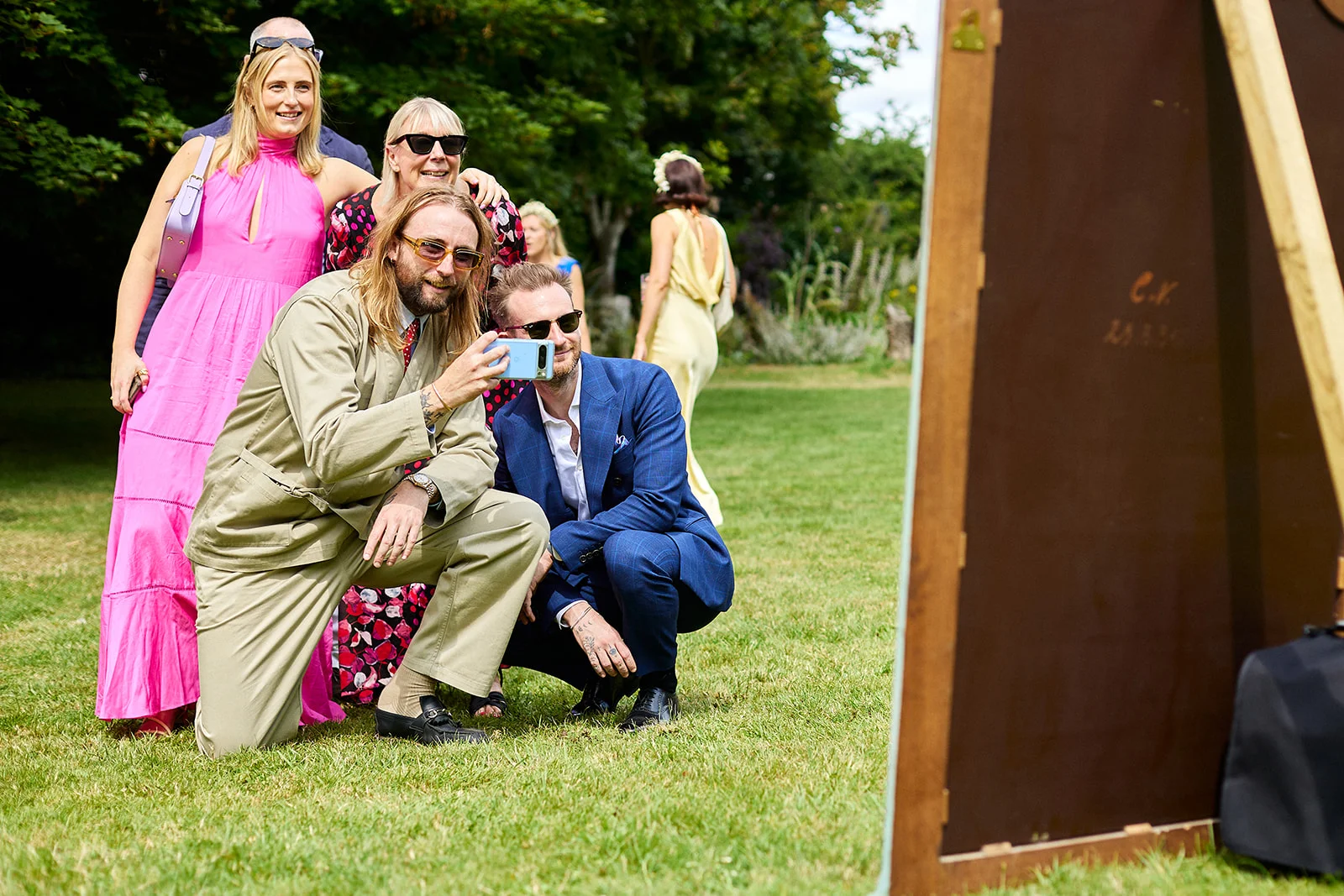 A group of wedding guests enjoying a sunny outdoor reception on the lawn, with two men crouching to take a selfie while other attendees gather around laughing.