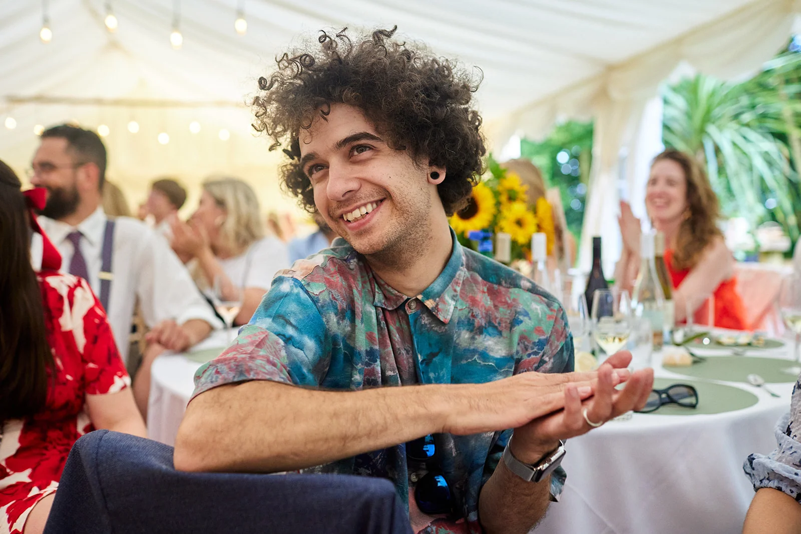 A smiling wedding guest with dark curly hair claps his hands at a table inside a brightly lit marquee during the reception.