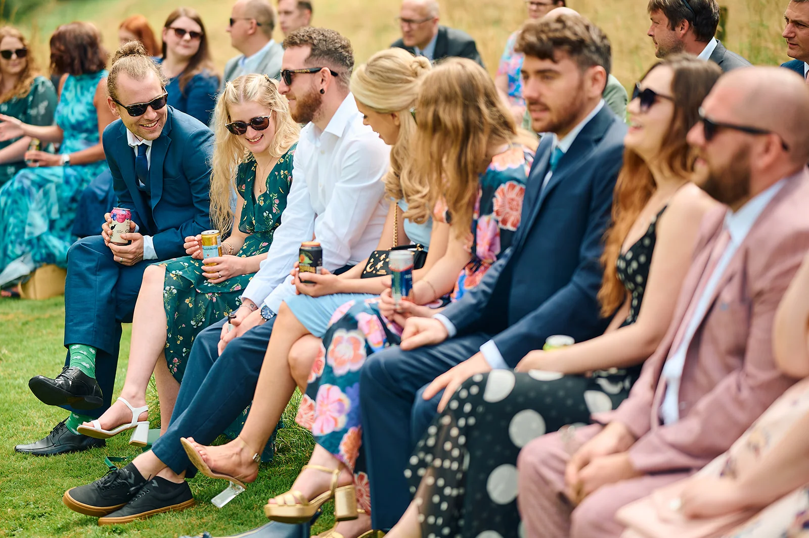A candid outdoor group shot of wedding guests laughing and chatting together on the grass during a sunny ceremony.