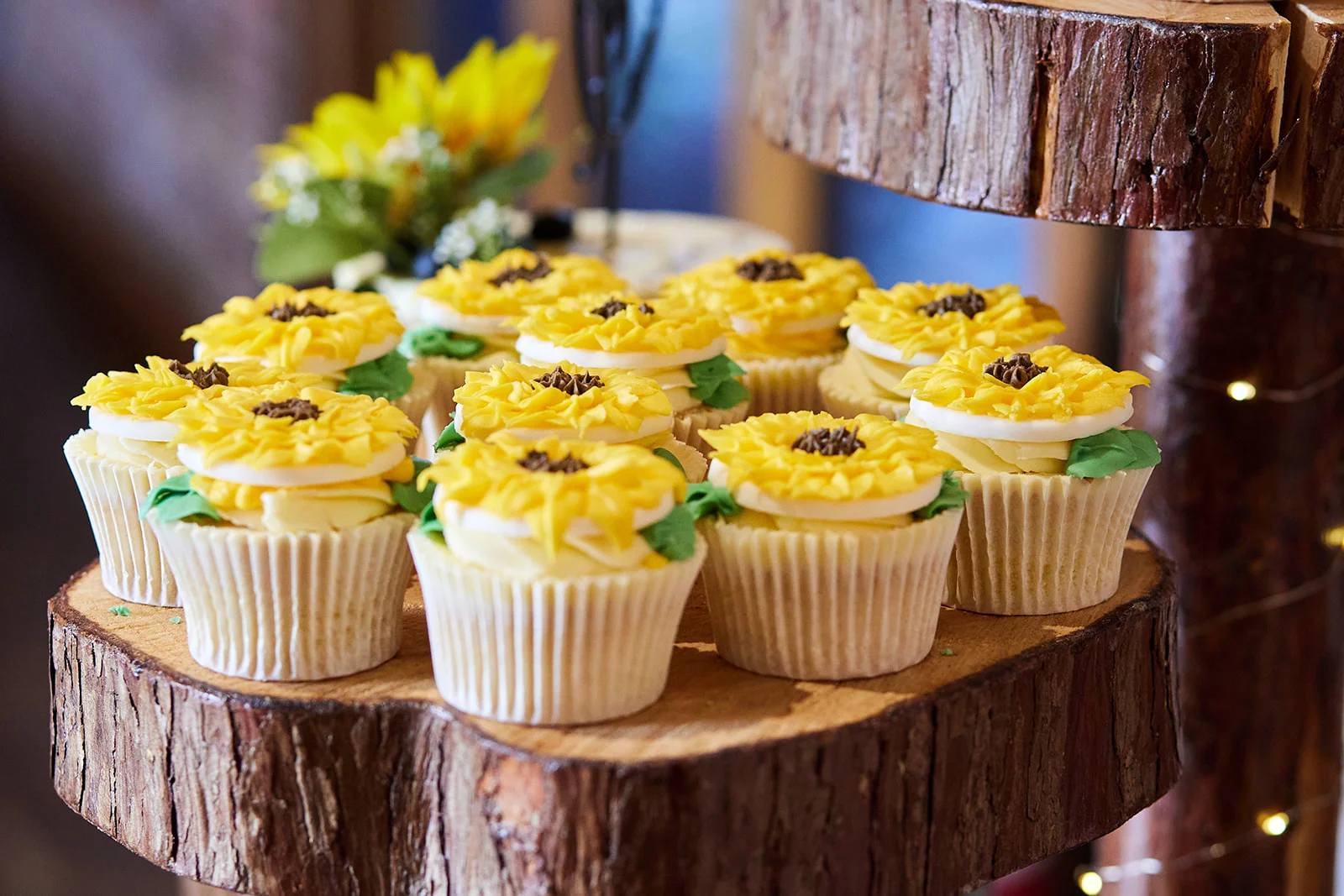 A selection of beautifully decorated sunflower-themed cupcakes displayed on a rustic wood slice tier at a wedding reception.