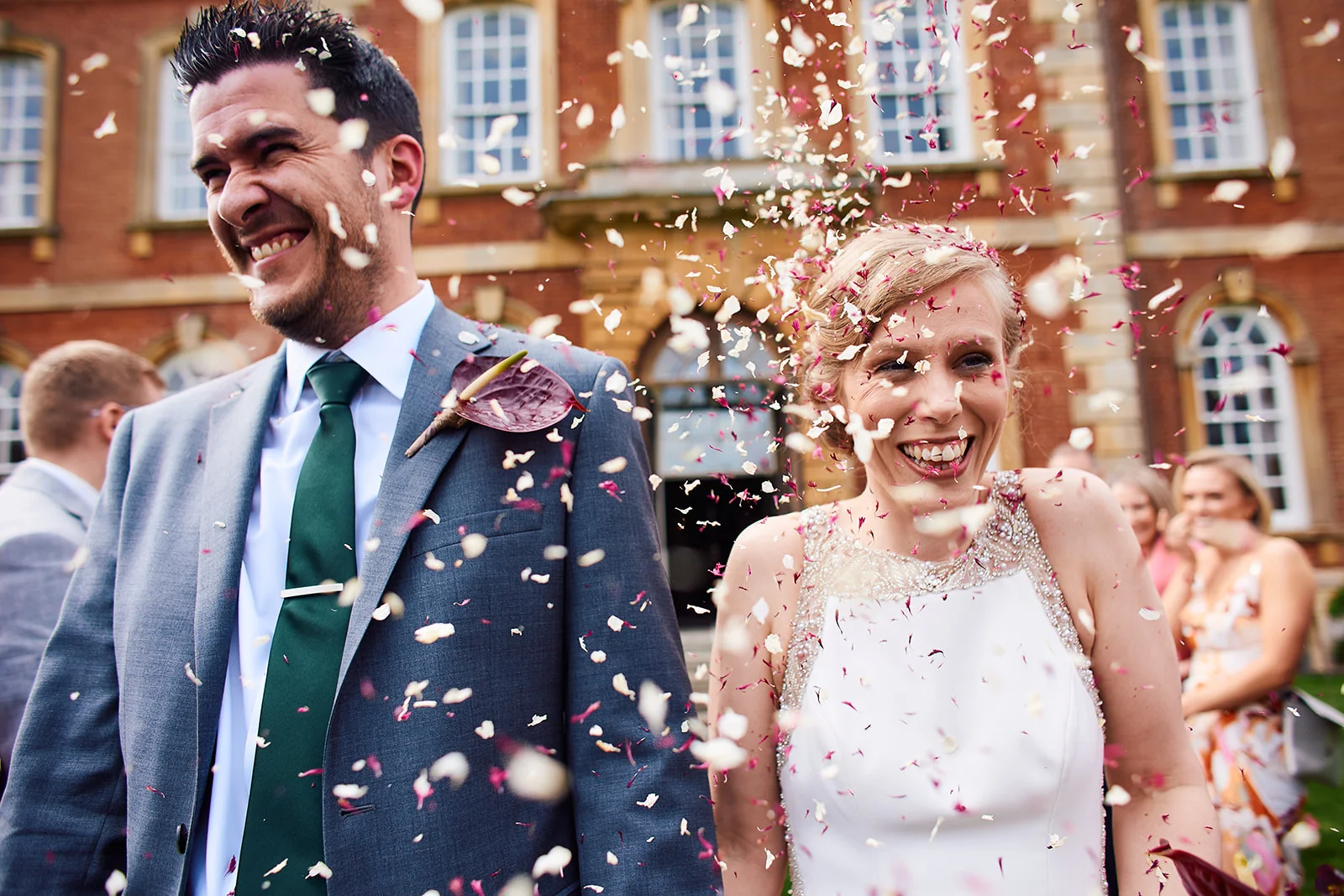 A joyful newlywed couple laughs as a cascade of mixed flower petal confetti falls around them outside a grand red brick country house.