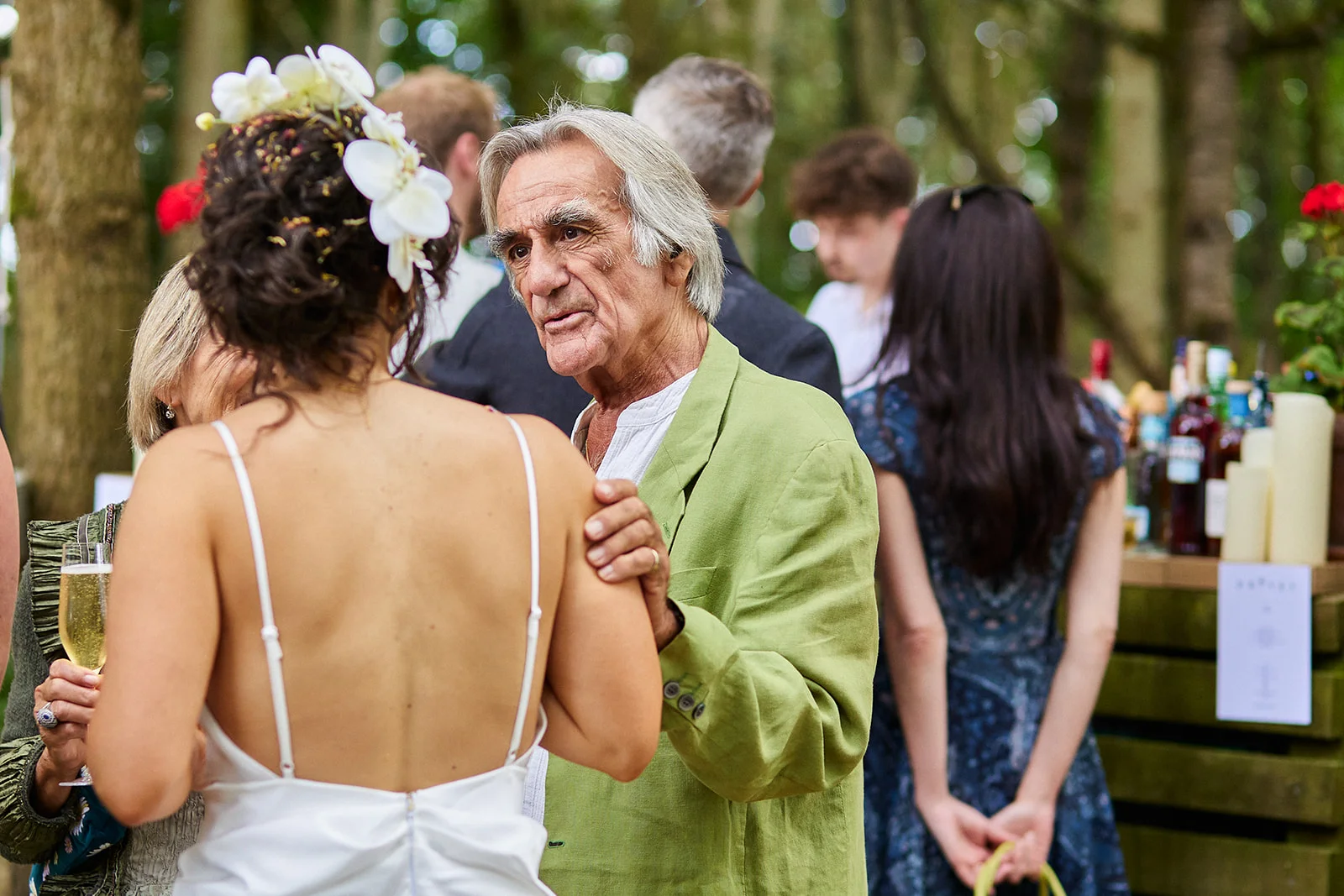 An older man in a green jacket places a hand gently on the shoulder of a bride wearing a white slip dress and white flowers in her hair during an outdoor wedding drinks reception.