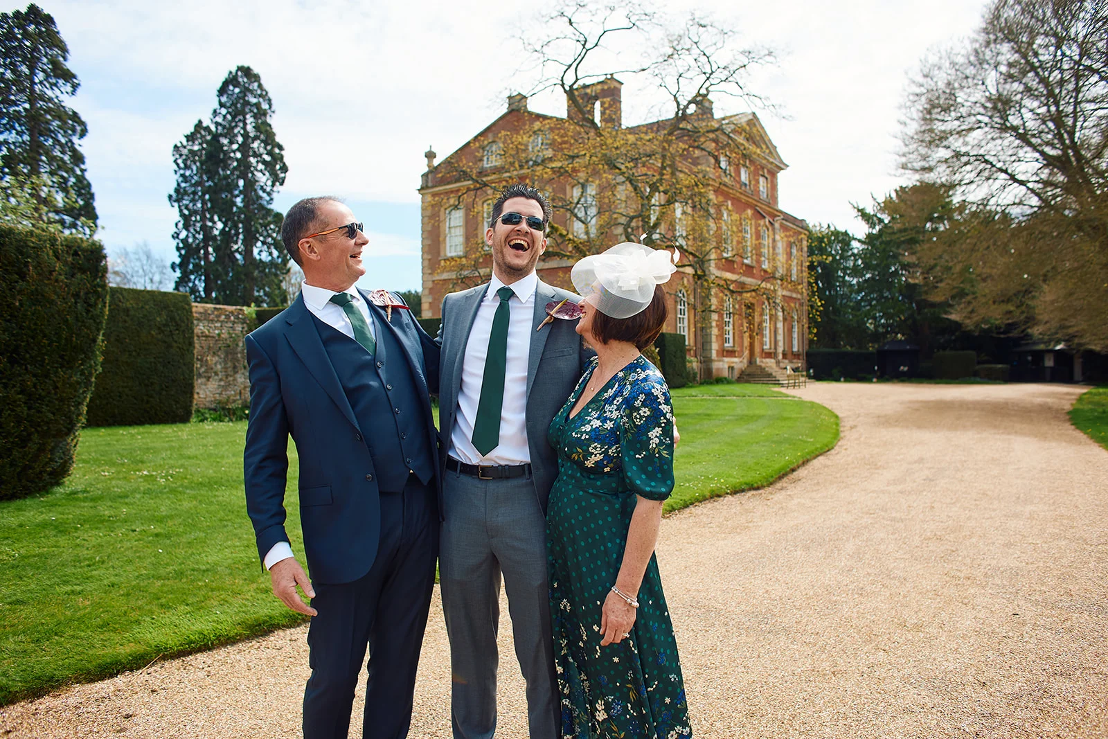 Three wedding guests laugh joyfully outdoors in front of a grand manor house, wearing a sharp suit and a floral dress with a fascinator.