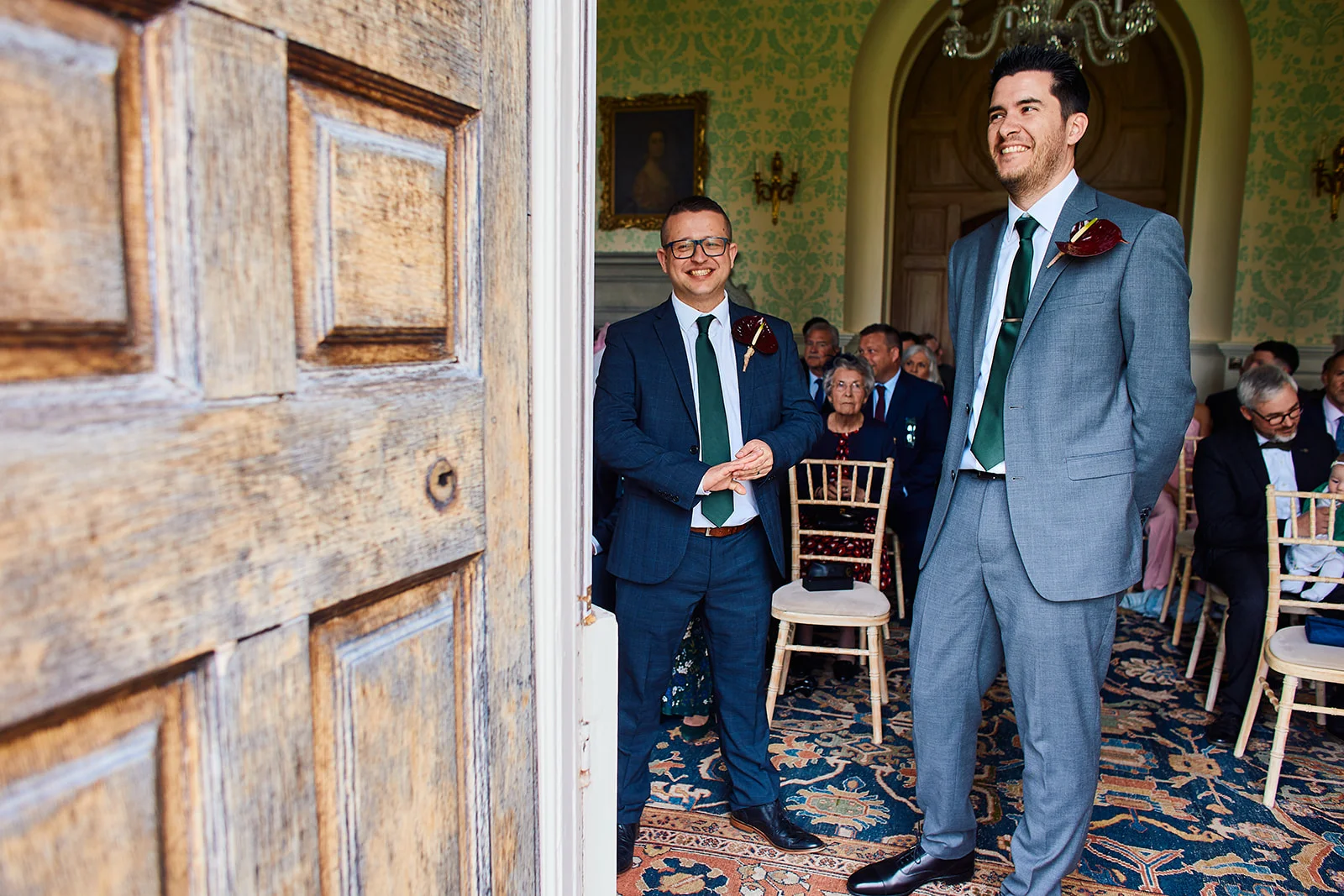 A smiling groom and groomsman wait inside a characterful venue with patterned wallpaper and ornate wood paneling, viewed through an open wooden doorway.
