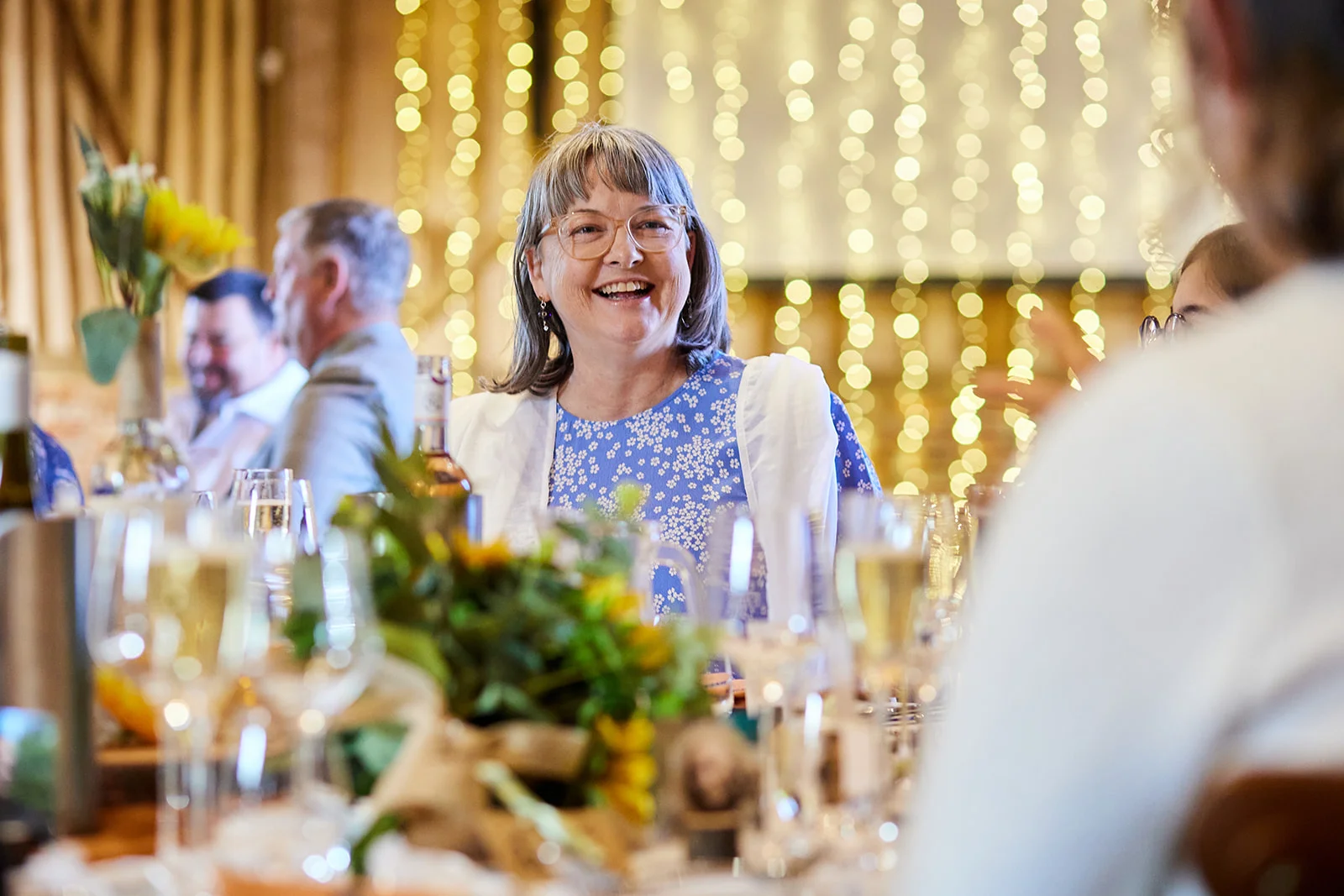 A smiling wedding guest laughs happily while seated at a festive banquet table illuminated by soft fairy lights.