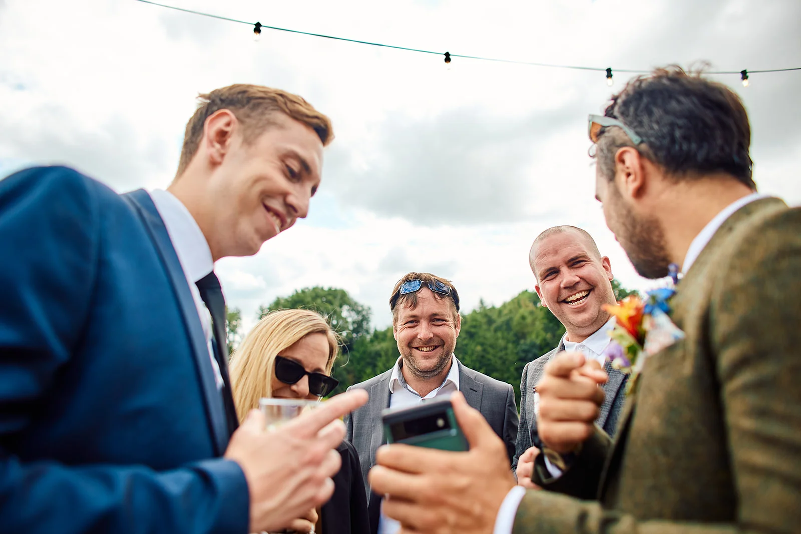 A group of wedding guests and groomsmen share a joyful, laughing moment outside while looking at a smartphone together under string lighting.