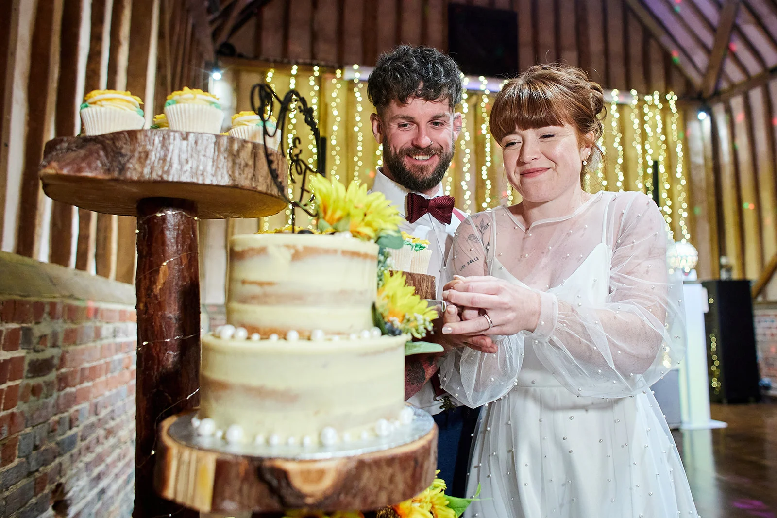 A smiling bride and groom cut into a rustic tiered wedding cake decorated with sunflowers inside a barn venue illuminated by fairy lights.