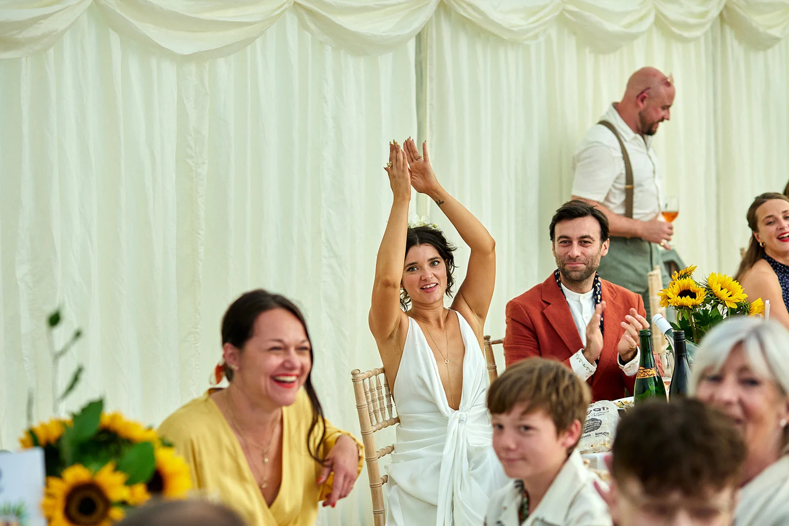 Wedding guests sit at long banquet tables inside a draped marquee, laughing and clapping enthusiastically during the speeches.