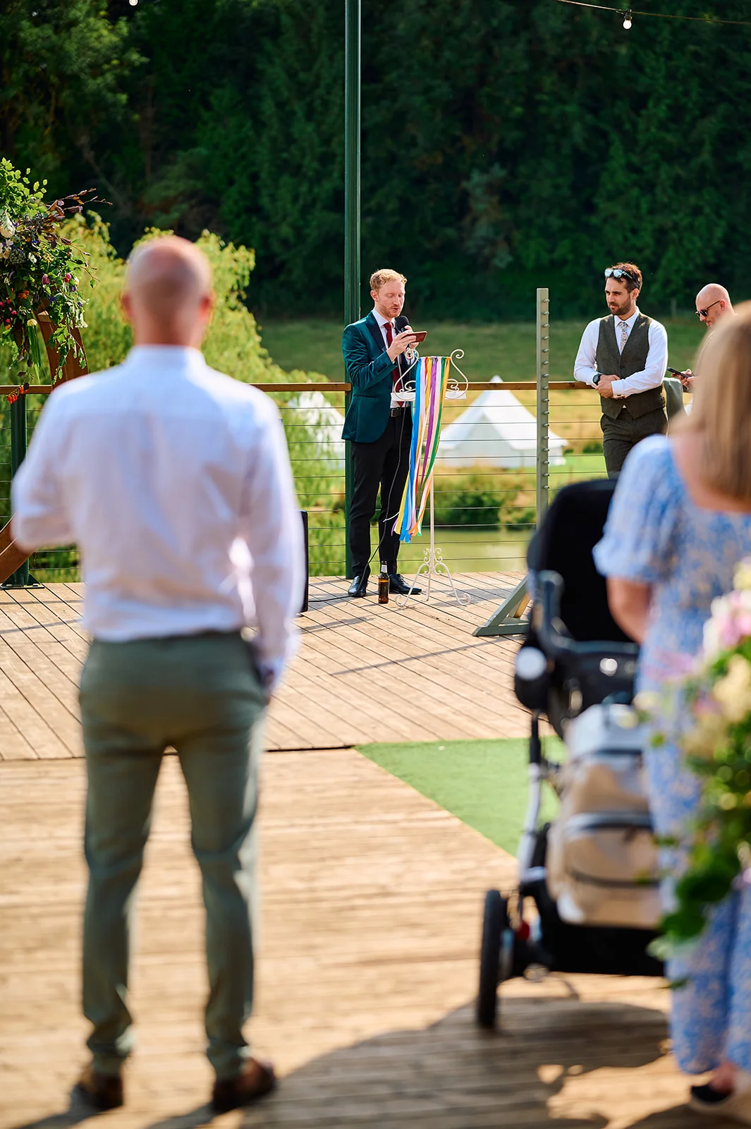 A groom delivers a speech outdoors on a wooden deck during a wedding reception, while guests watch and listen.