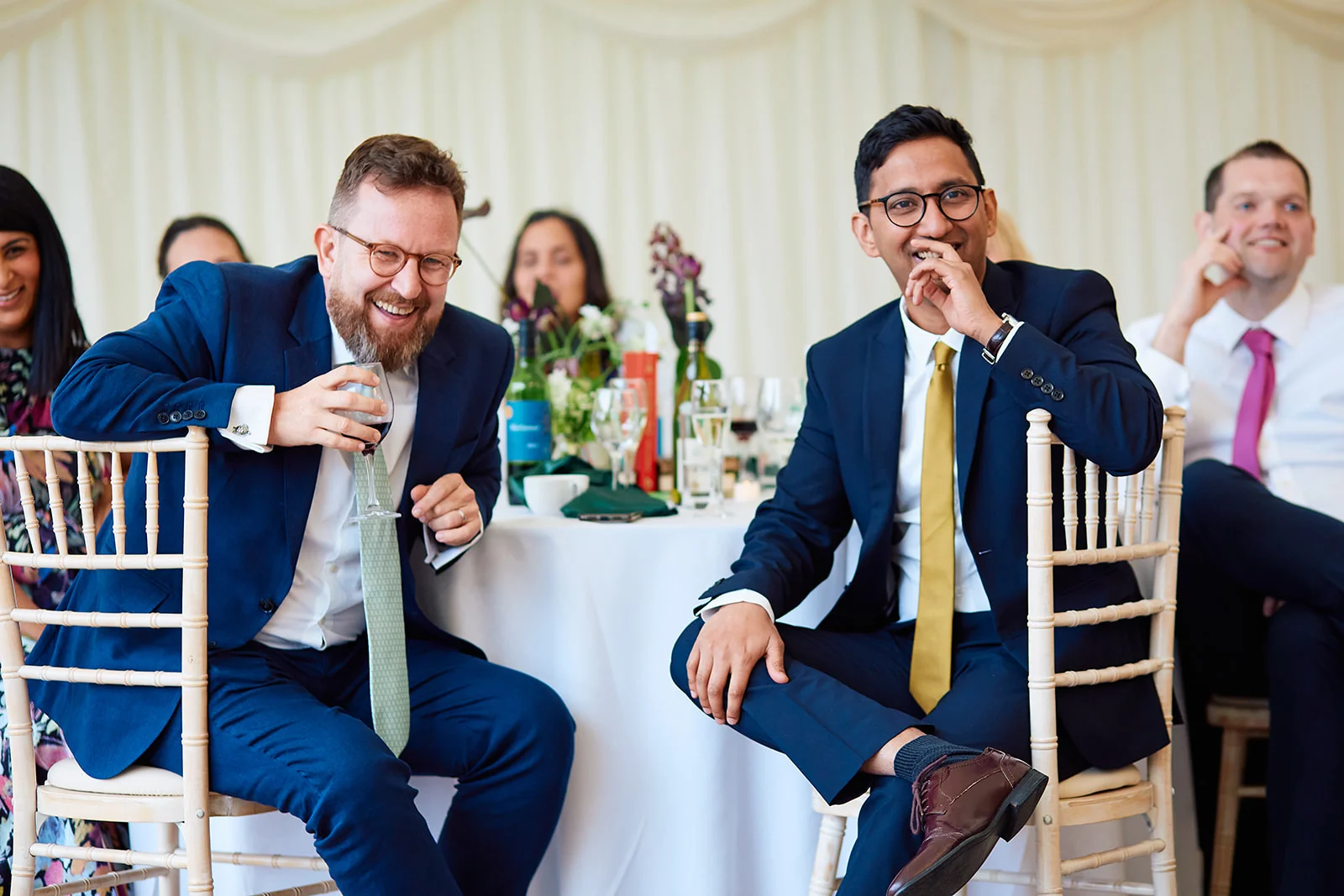 Two wedding guests in navy suits and ties sit at a banquet table inside a marquee, laughing heartily during the wedding speeches.