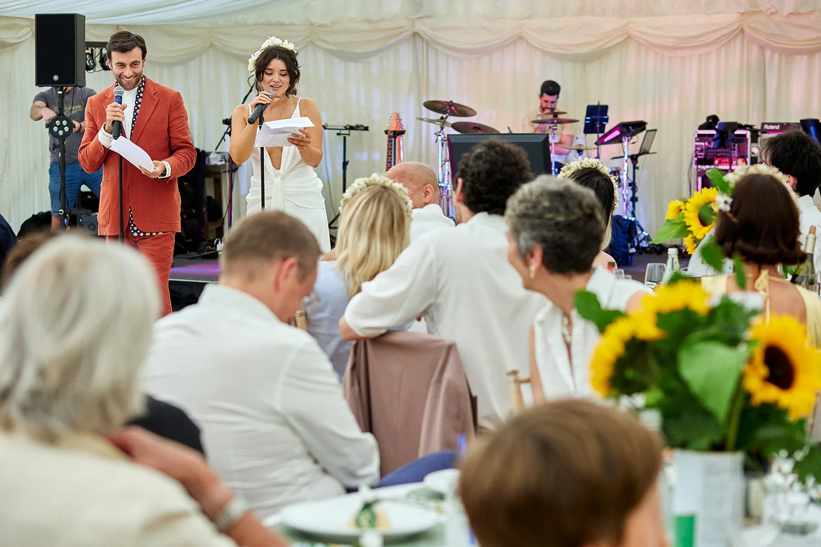 A bride and groom deliver speeches into microphones inside a brightly lit marquee during their wedding reception, while seated guests look on.