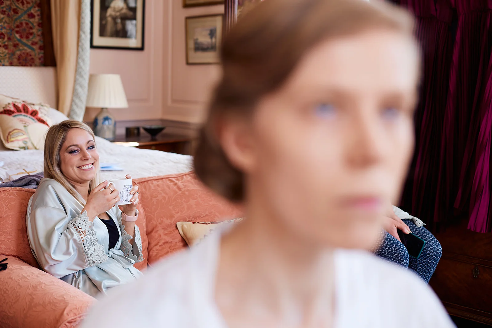 A bridesmaid smiles while holding a mug and relaxing on a coral sofa in a bedroom while another person sits softly blurred in the foreground during morning preparations.