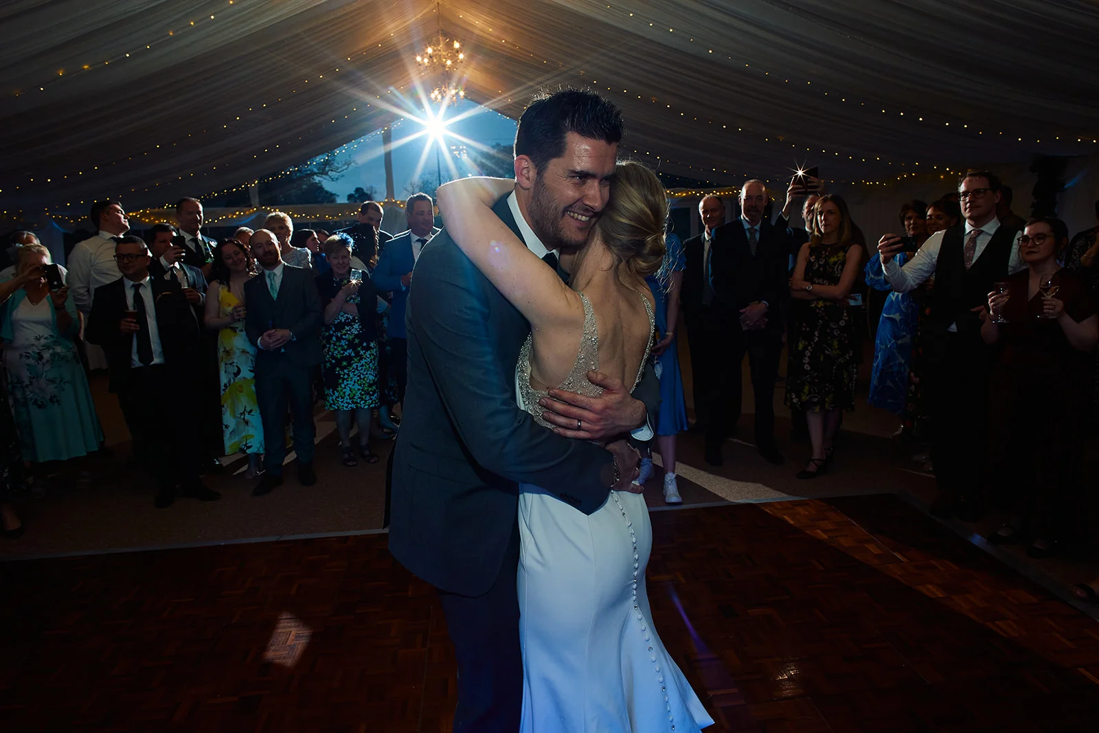The newlywed couple shares their first dance on a wooden dance floor inside a marquee illuminated by fairy lights, while guests look on and cheer.