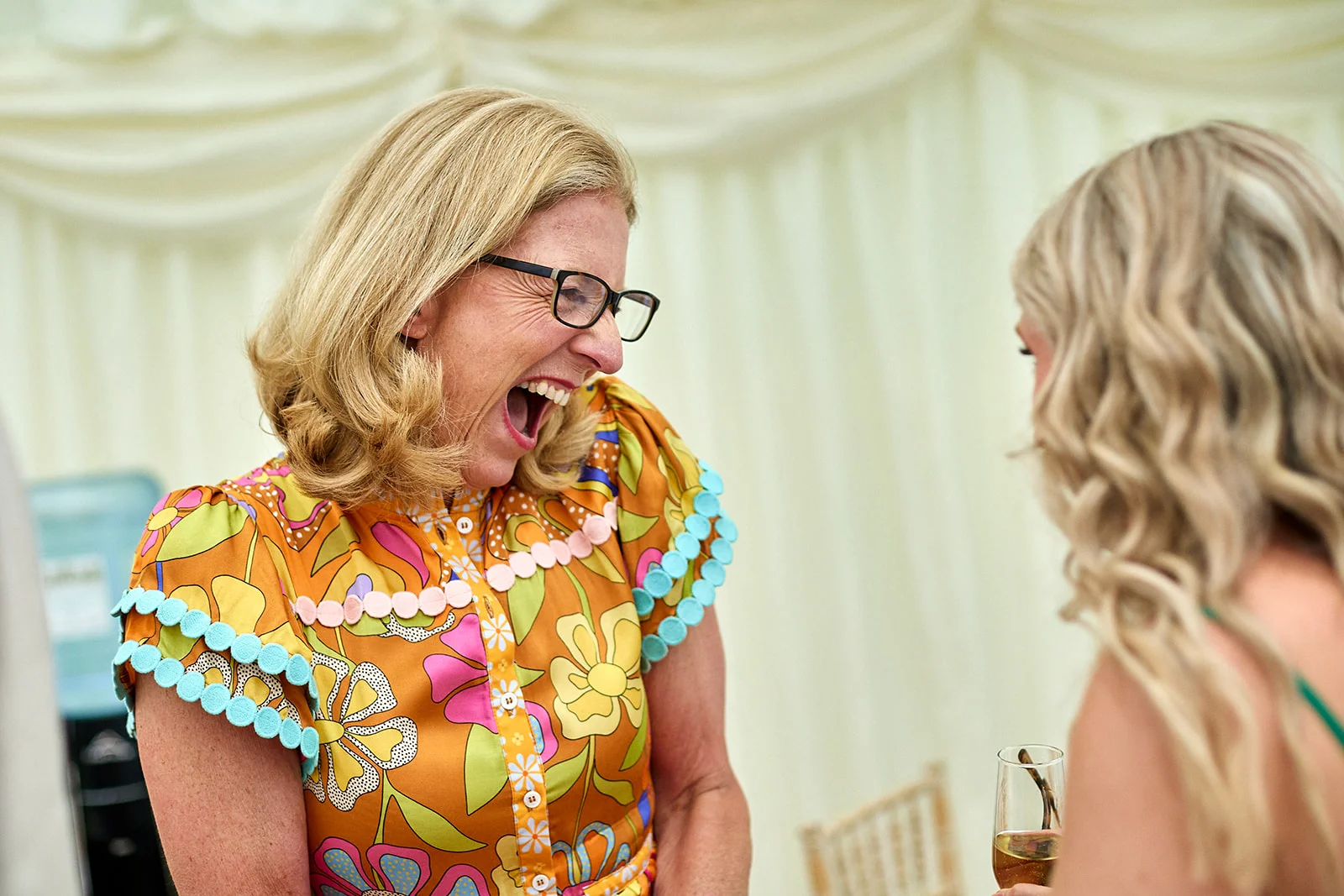 A wedding guest in a vibrant floral print dress laughs joyfully while interacting with another guest holding a glass inside a draped marquee.