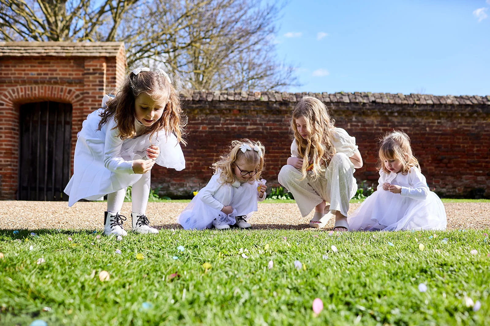 Four young flower girls in white dresses collect colourful confetti from a lush green lawn outside a historic brick building.