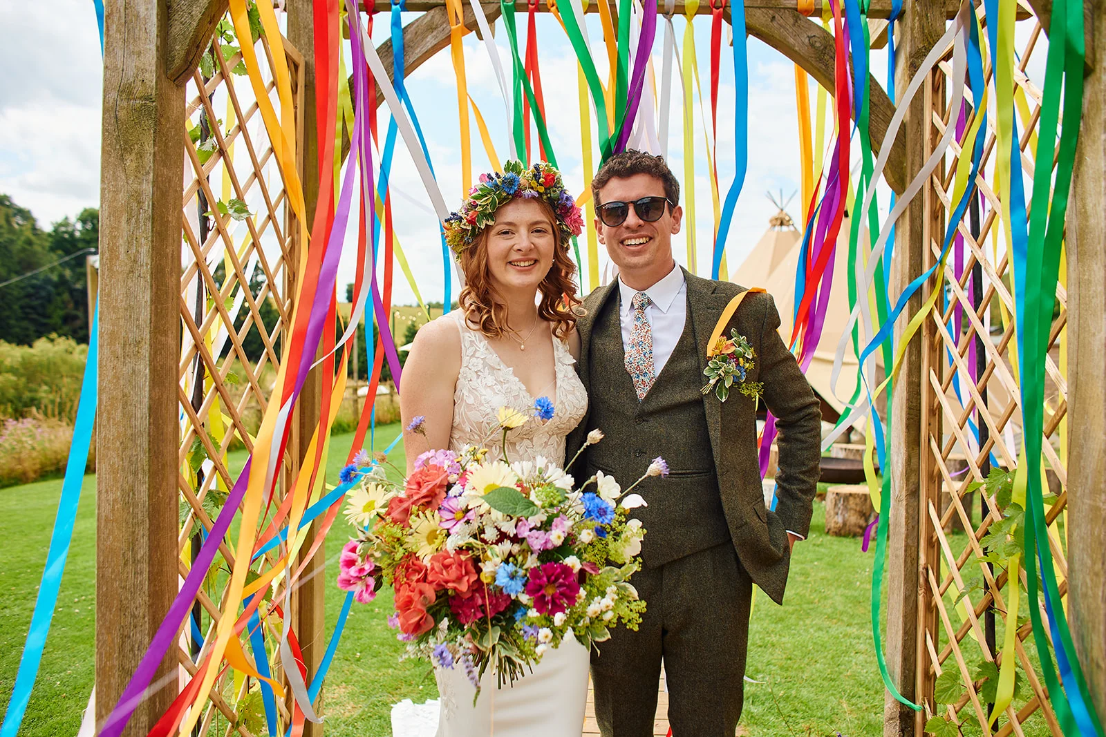 A smiling bride wearing a flower crown and holding a colourful wildflower bouquet stands with the groom in a tweed suit beneath a wooden arch decorated with vibrant hanging ribbons.