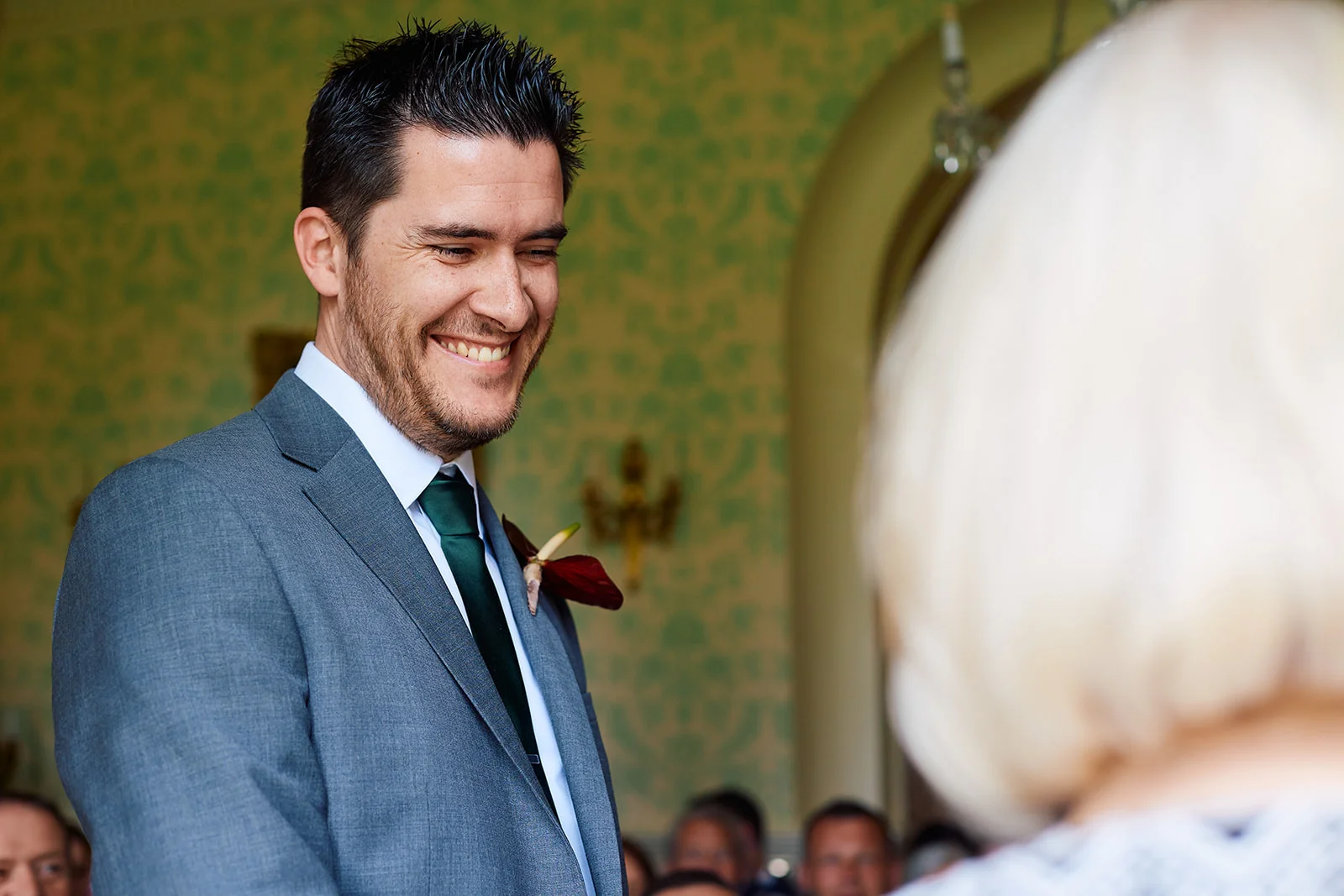 A smiling groom in a grey suit and green tie looks lovingly toward his partner during the wedding ceremony.