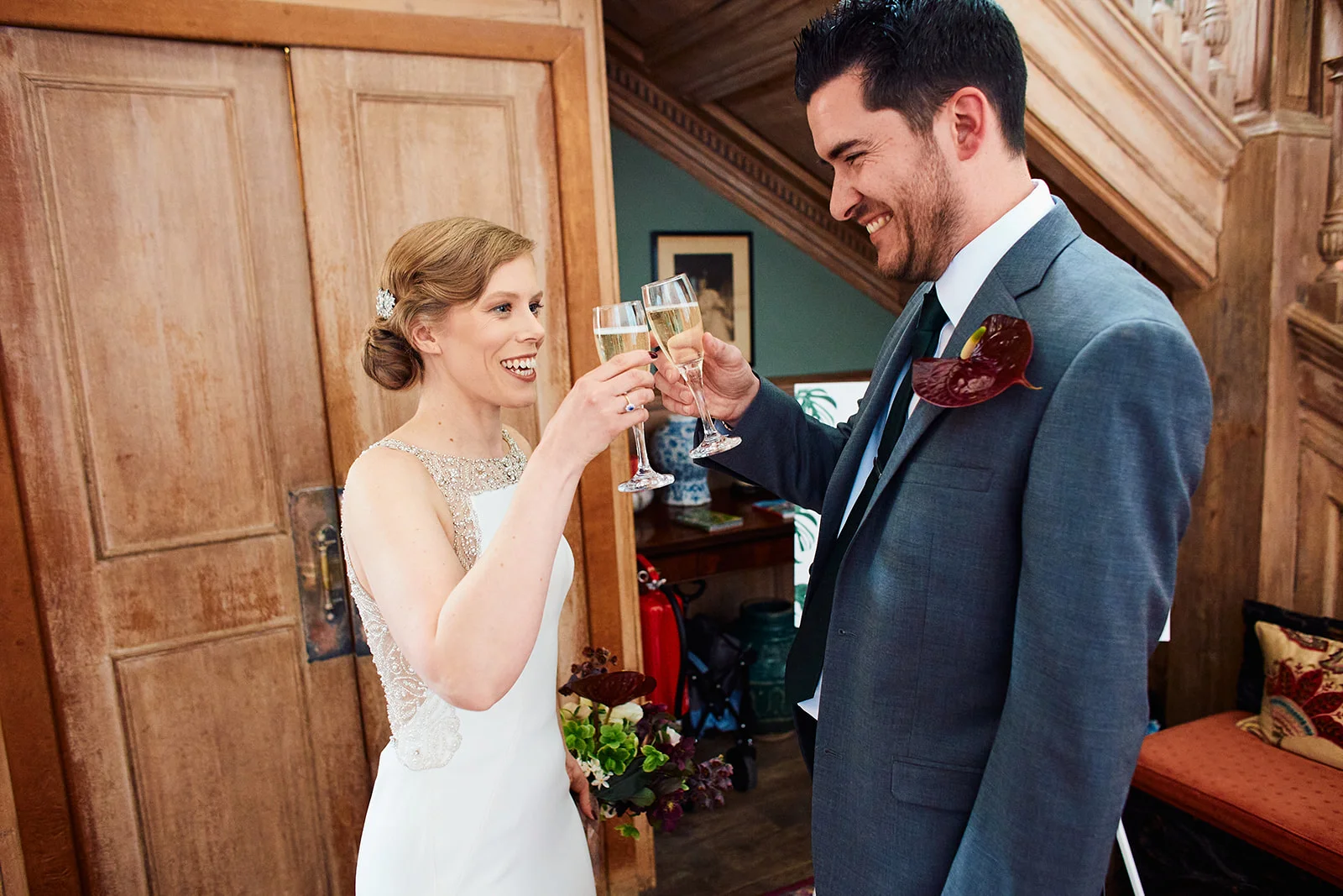 A smiling bride and groom joyfully toast with champagne flutes near wooden panelled doors inside a classic venue.