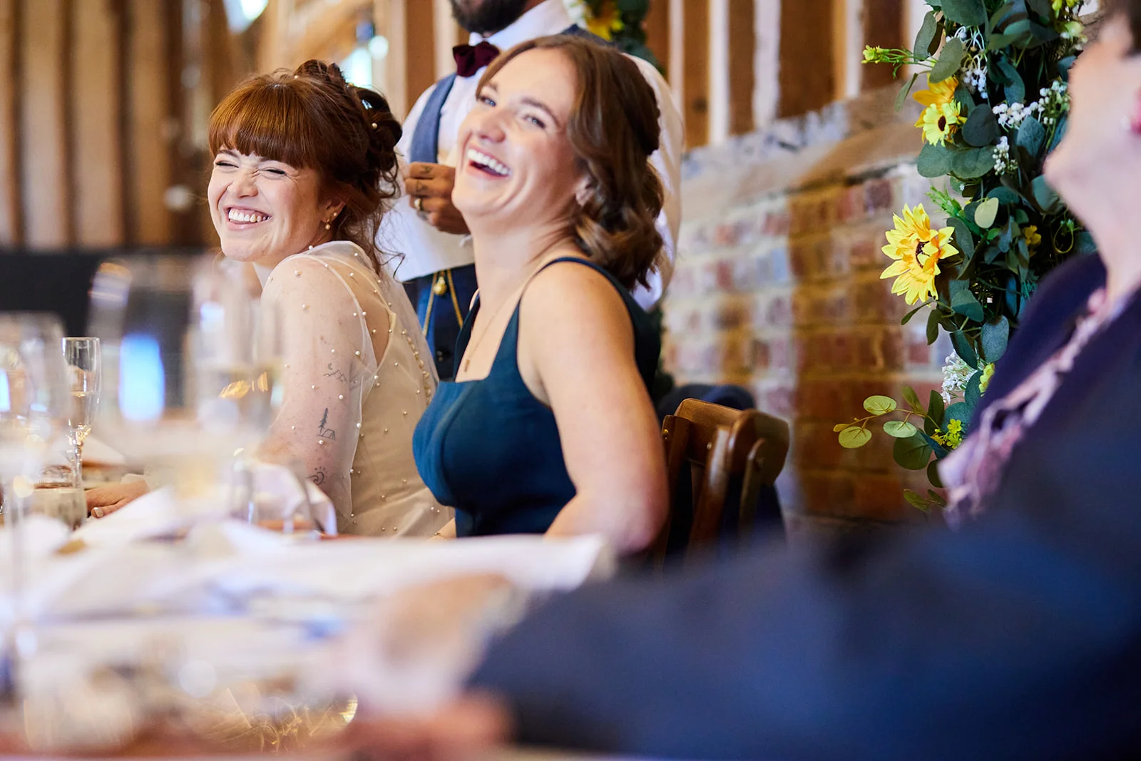 Two smiling brides share a joyful, laughing moment together at the top table during wedding breakfast speeches in a rustic barn venue decorated with sunflowers.