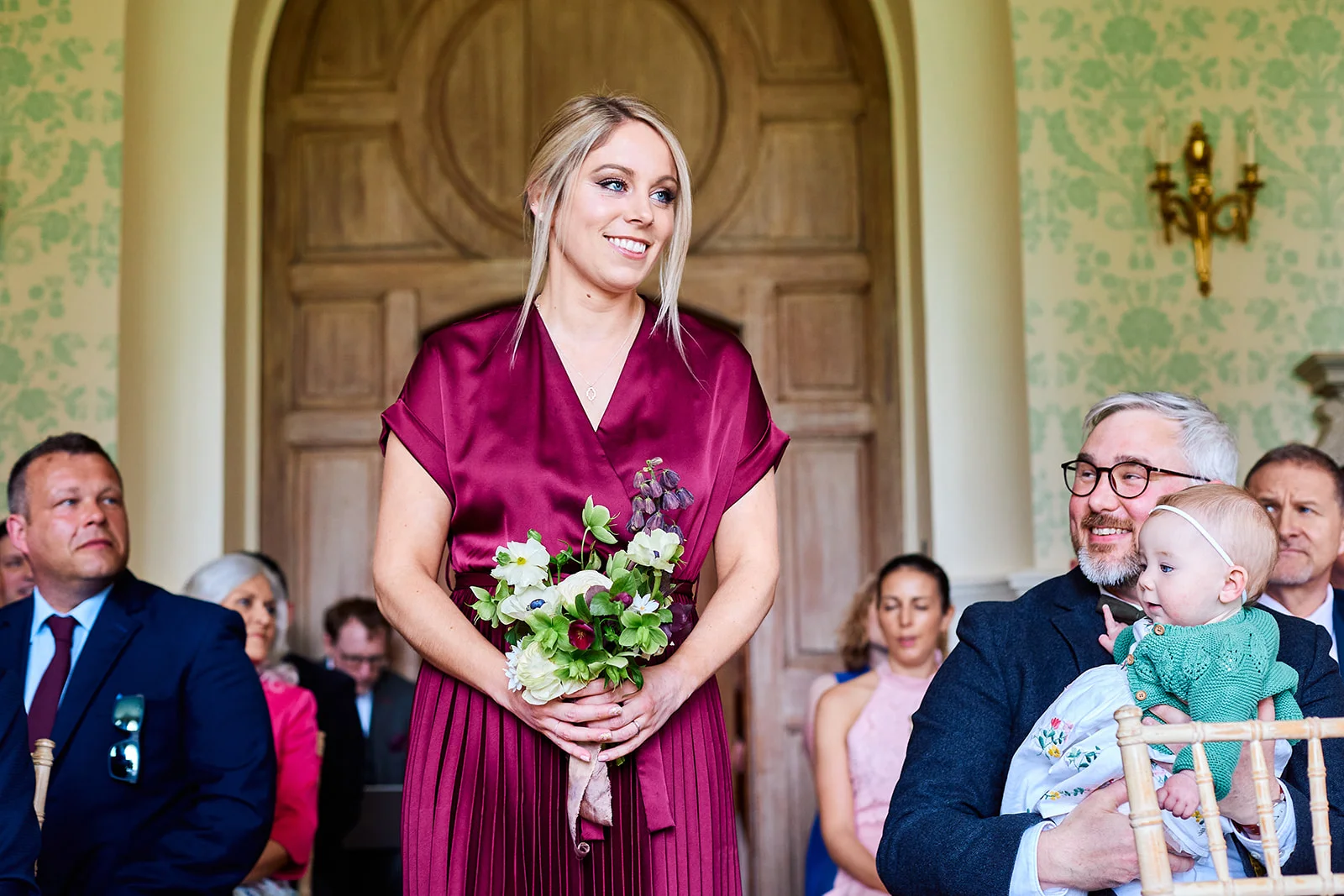 A smiling bridesmaid in a burgundy dress holding a bouquet stands before seated guests inside an elegant country house.