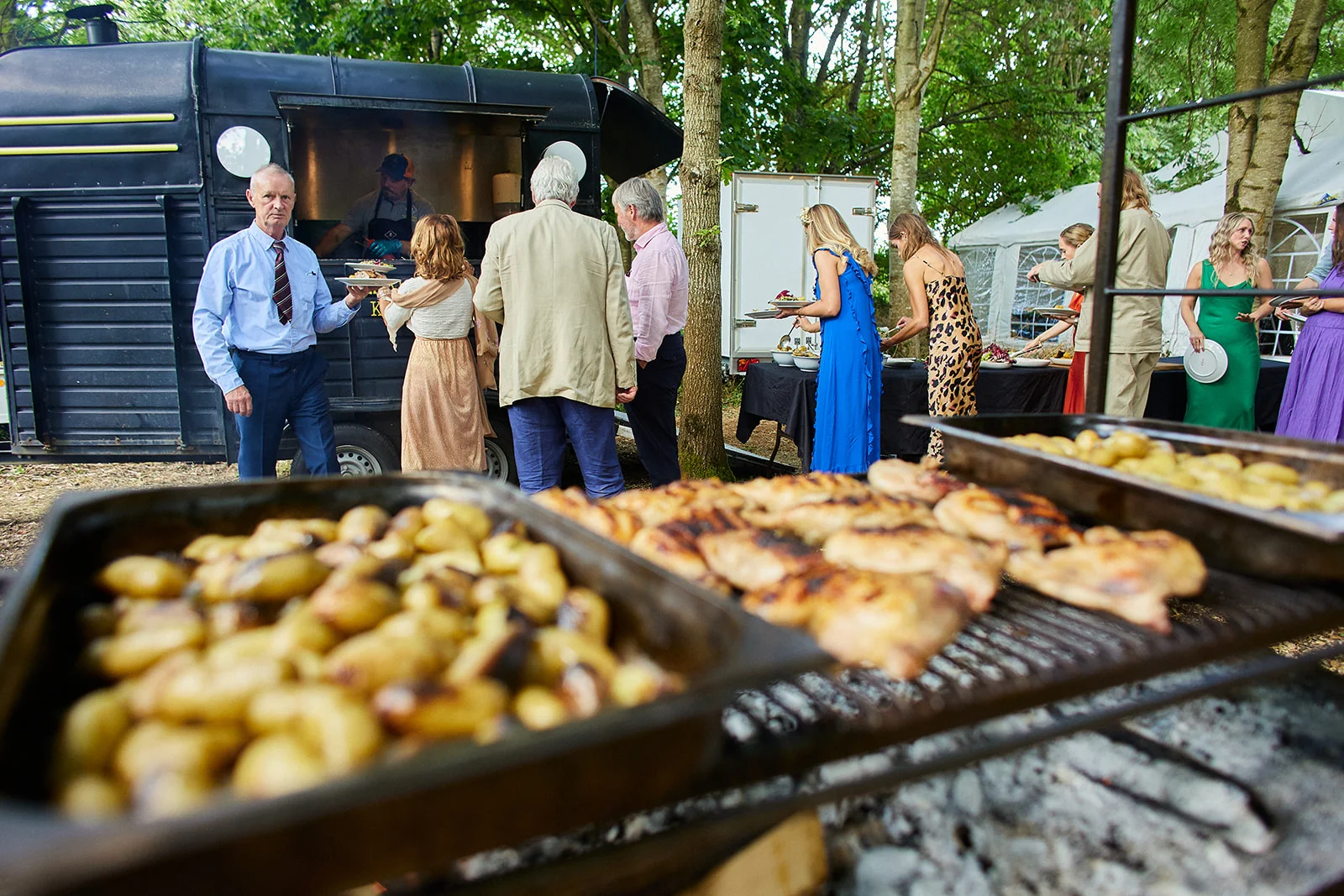 Guests queue at a converted horsebox food truck for a barbecue wedding feast in a woodland setting.