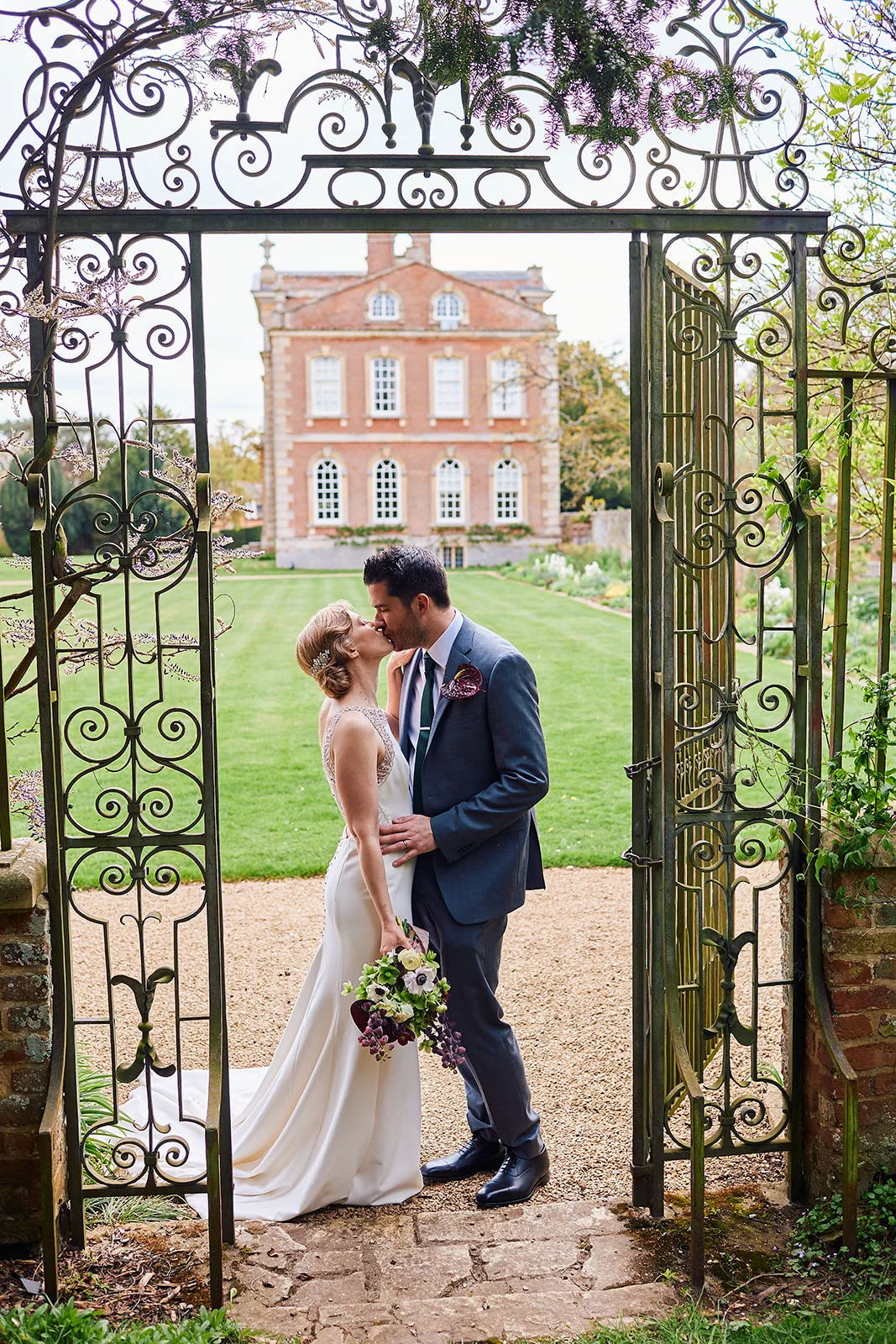 A bride and groom share a tender kiss framed by an ornate wrought iron garden gate with a grand country house in the background.