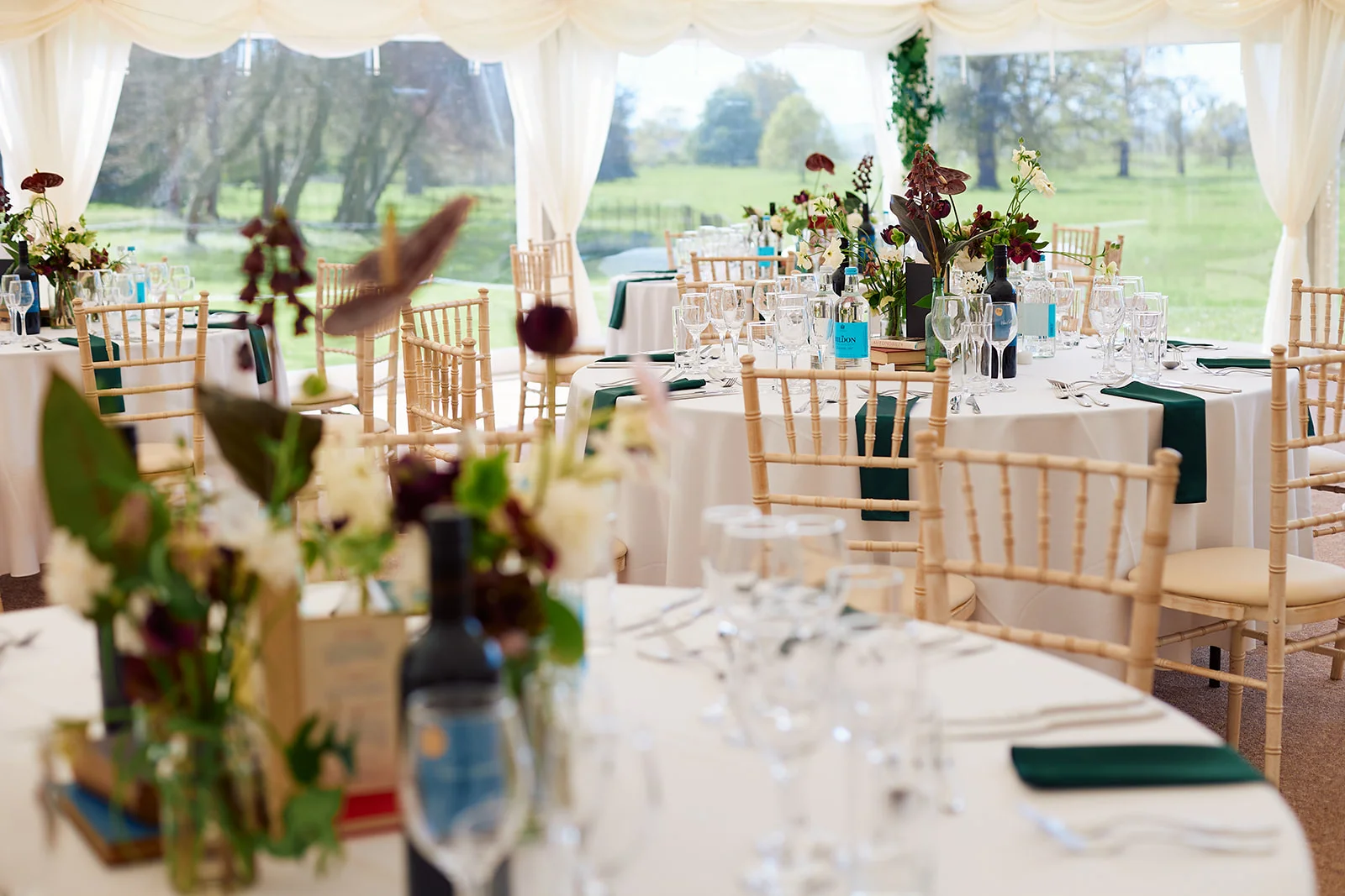 A beautifully styled marquee wedding reception featuring round tables with crisp white linens, green napkins, and elegant floral centrepieces with scenic views outside.