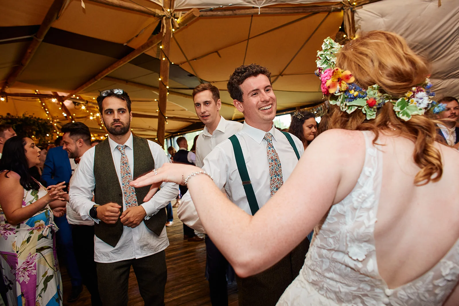 A bride wearing a floral crown and lace gown dances with her smiling groom inside a brightly lit sailcloth marquee filled with guests.