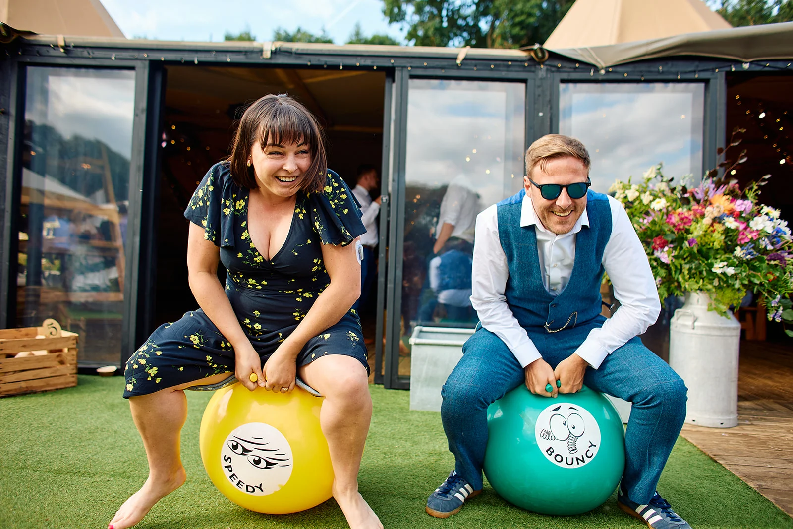 A joyful couple laugh while bouncing on space hoppers on the lawn outside a marquee venue on a bright summer wedding day.