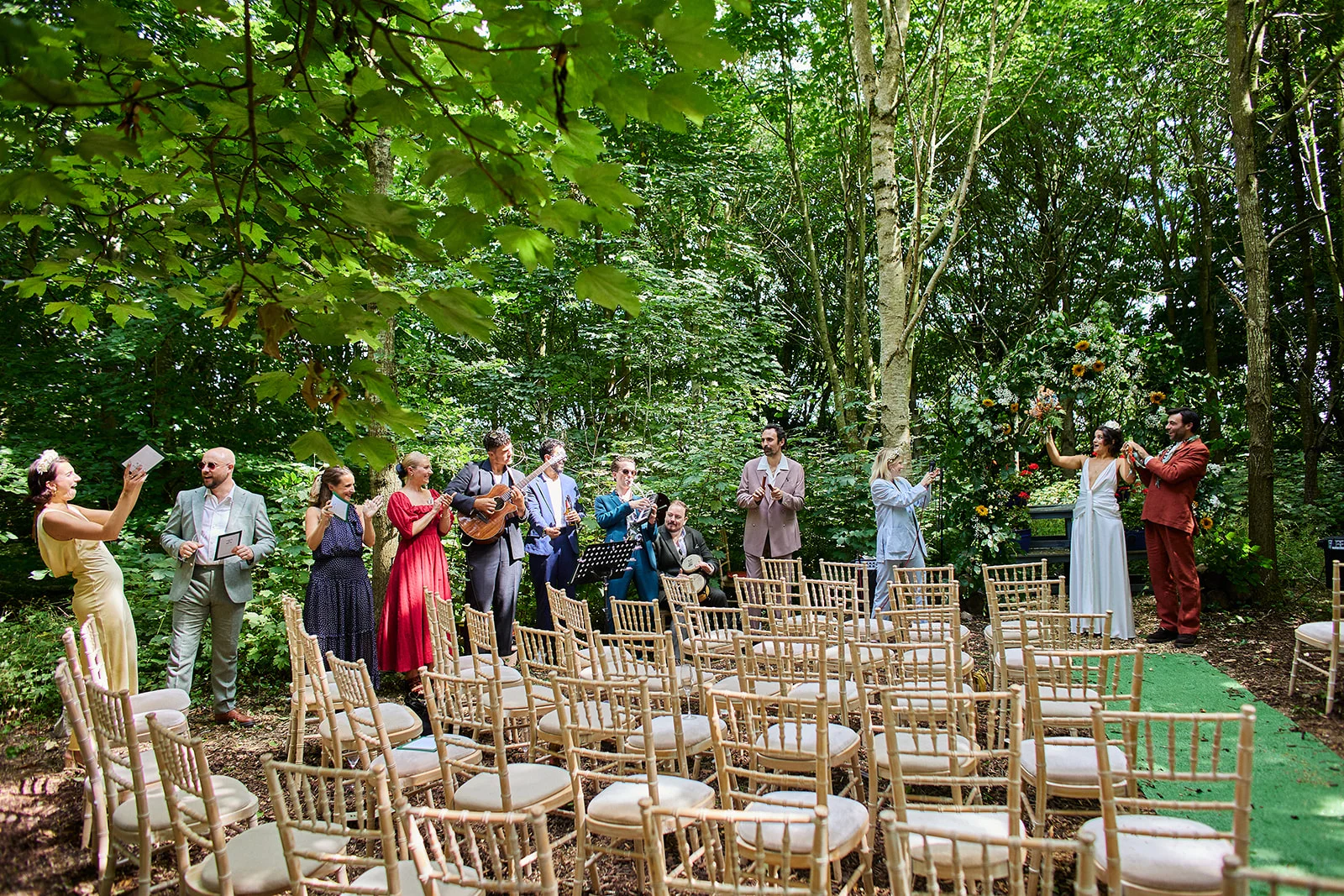 Guests stand and applaud in a sunlit woodland clearing during an outdoor wedding ceremony, with musicians playing nearby.