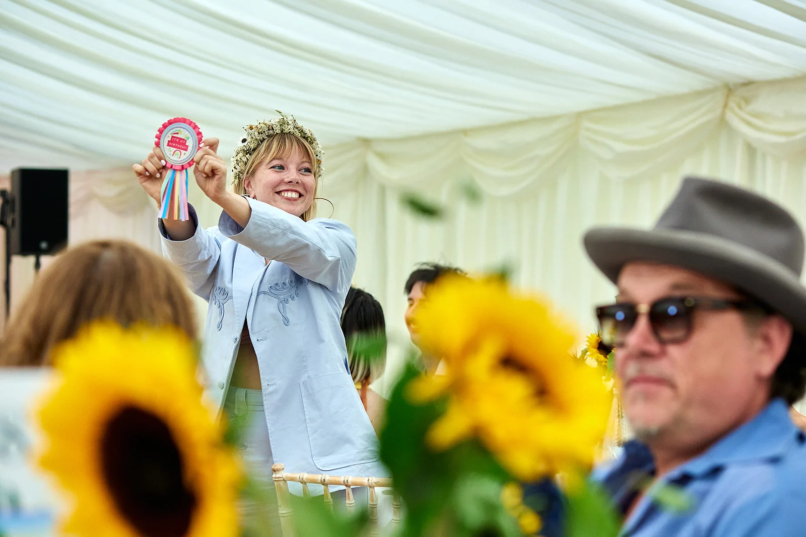 A joyful wedding guest wearing a floral crown and light blue suit cheerfully holds up a rosette inside a marquee decorated with bright sunflowers.